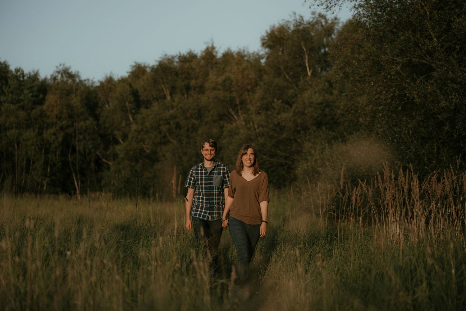 Couple holding hands walking through a sunlit meadow with tall grass and lush green trees in the background.