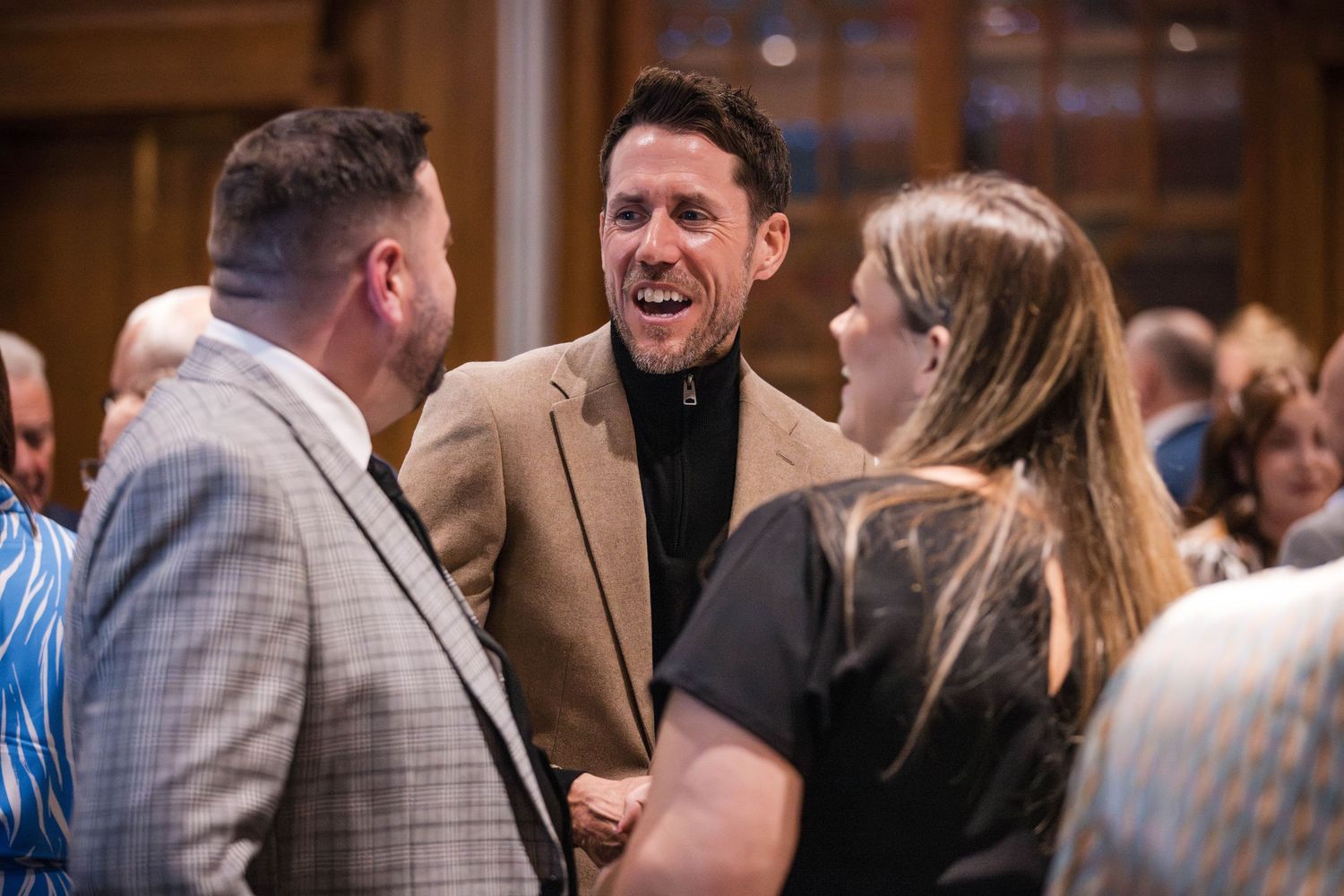 People laughing and socializing at an indoor event, with one wearing a tan blazer and another in a gray suit.