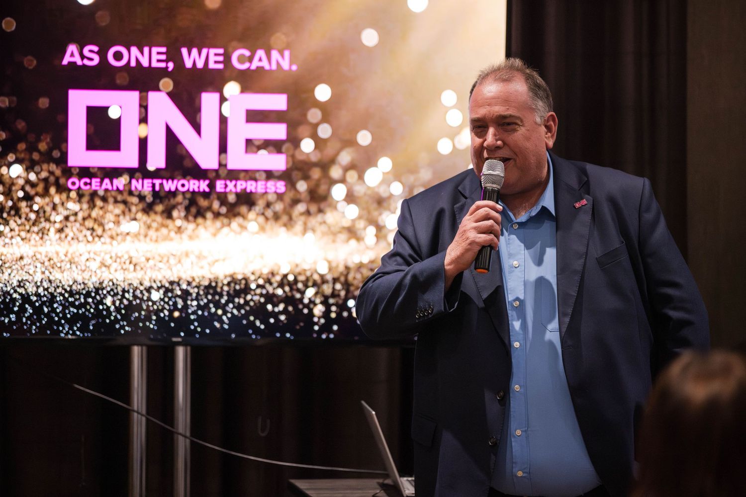 A person wearing a dark suit speaks on stage with bokeh lights and glowing effects in the background.