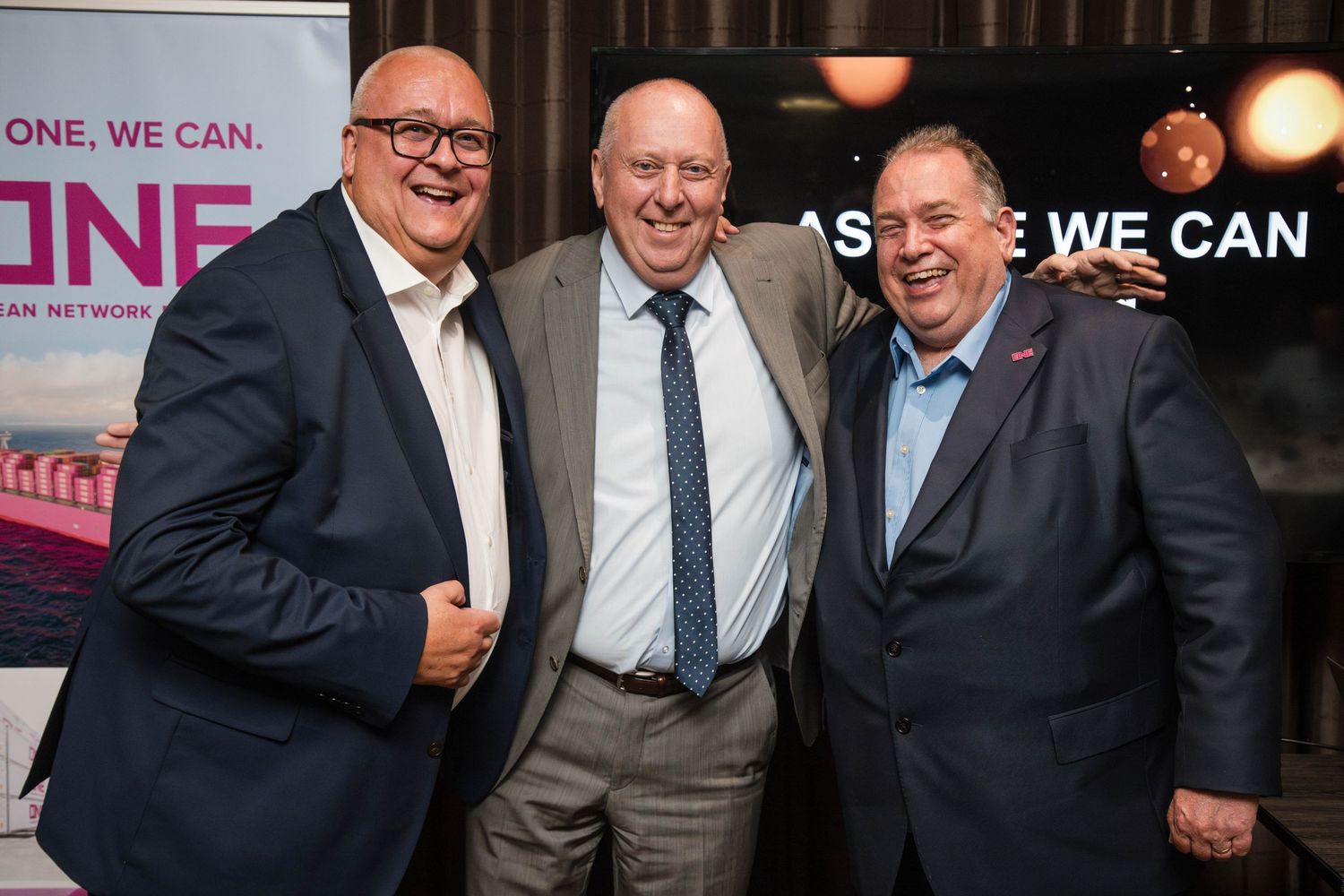 Three men in business attire pose together at an event with a backdrop displaying 'WE CAN' signage.