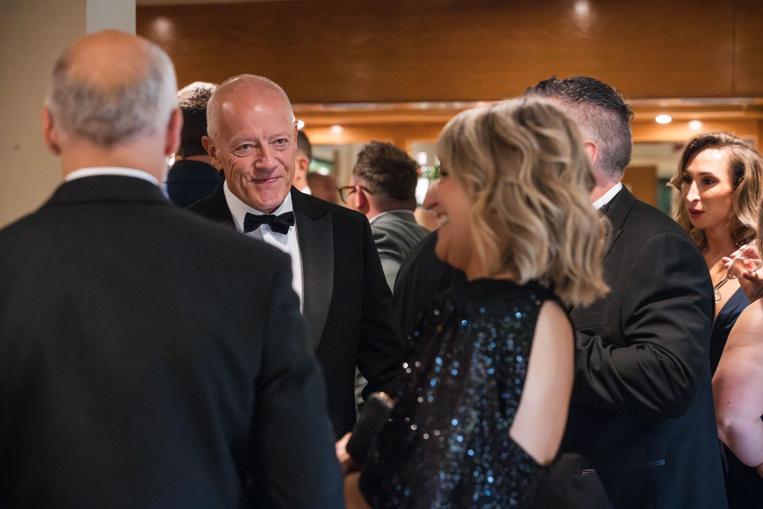 People in formal attire socializing at an elegant event with someone wearing a black tuxedo and another in a sparkling dress.