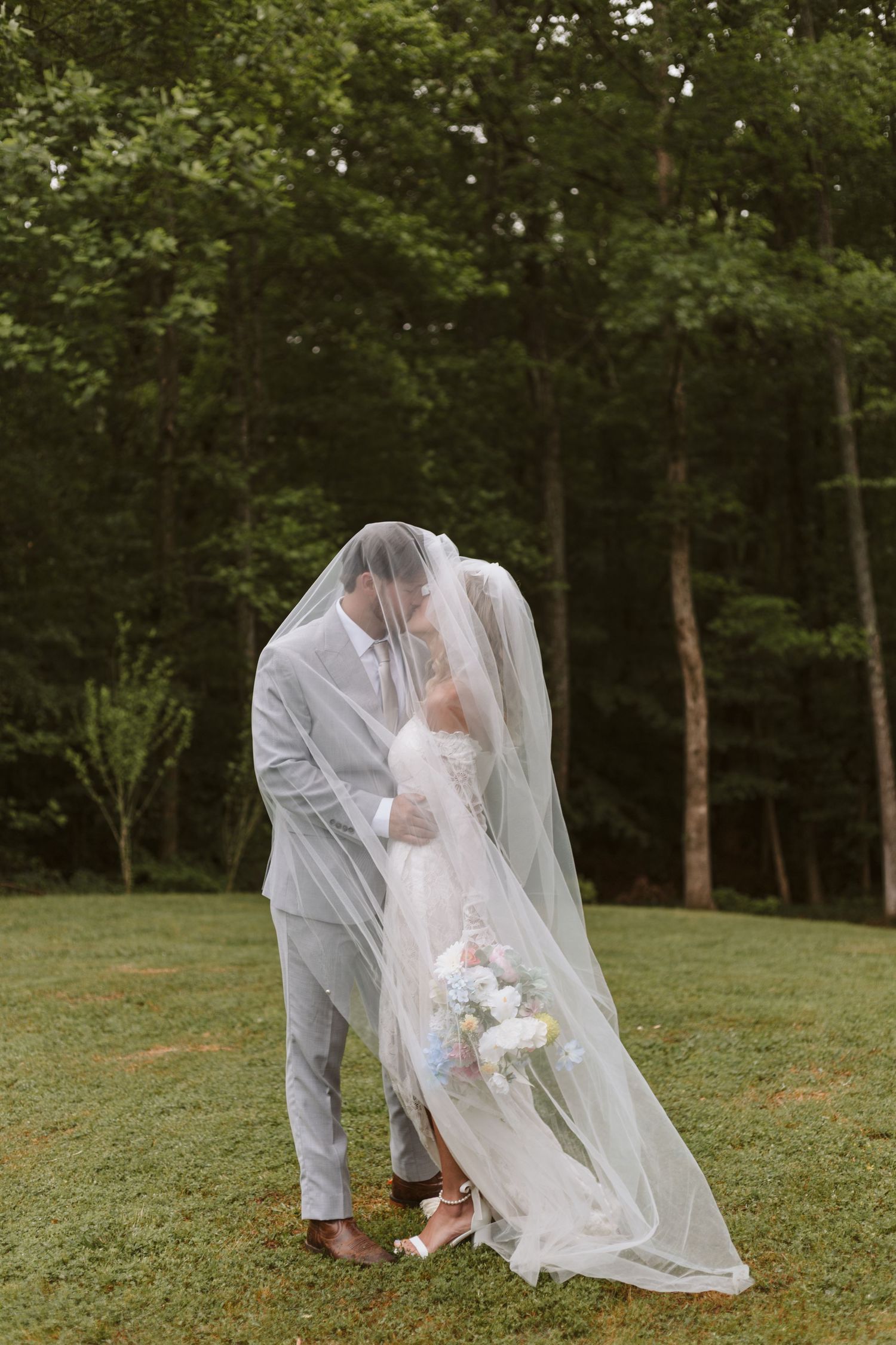 A bride and groom sharing an embrace under a white vail in a wooded field