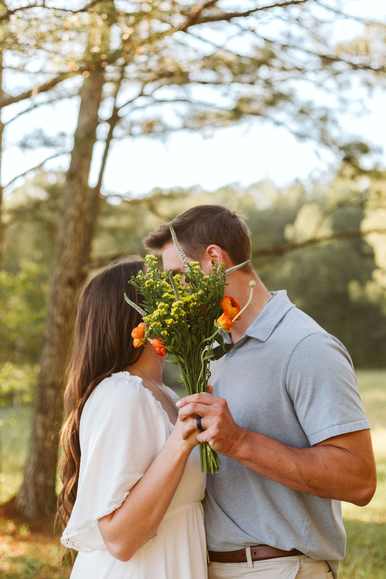 Couple sharing a kiss while holding a bouquet of flowers in front of their faces near a wooded area