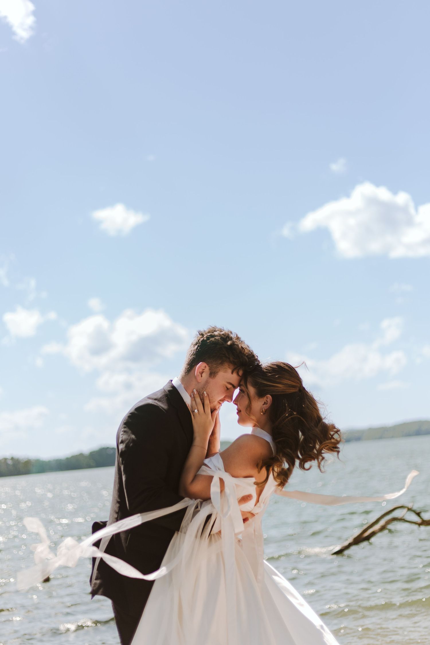 Bride holding the grooms face in a tender embrace with a white flowy dress on a lake.