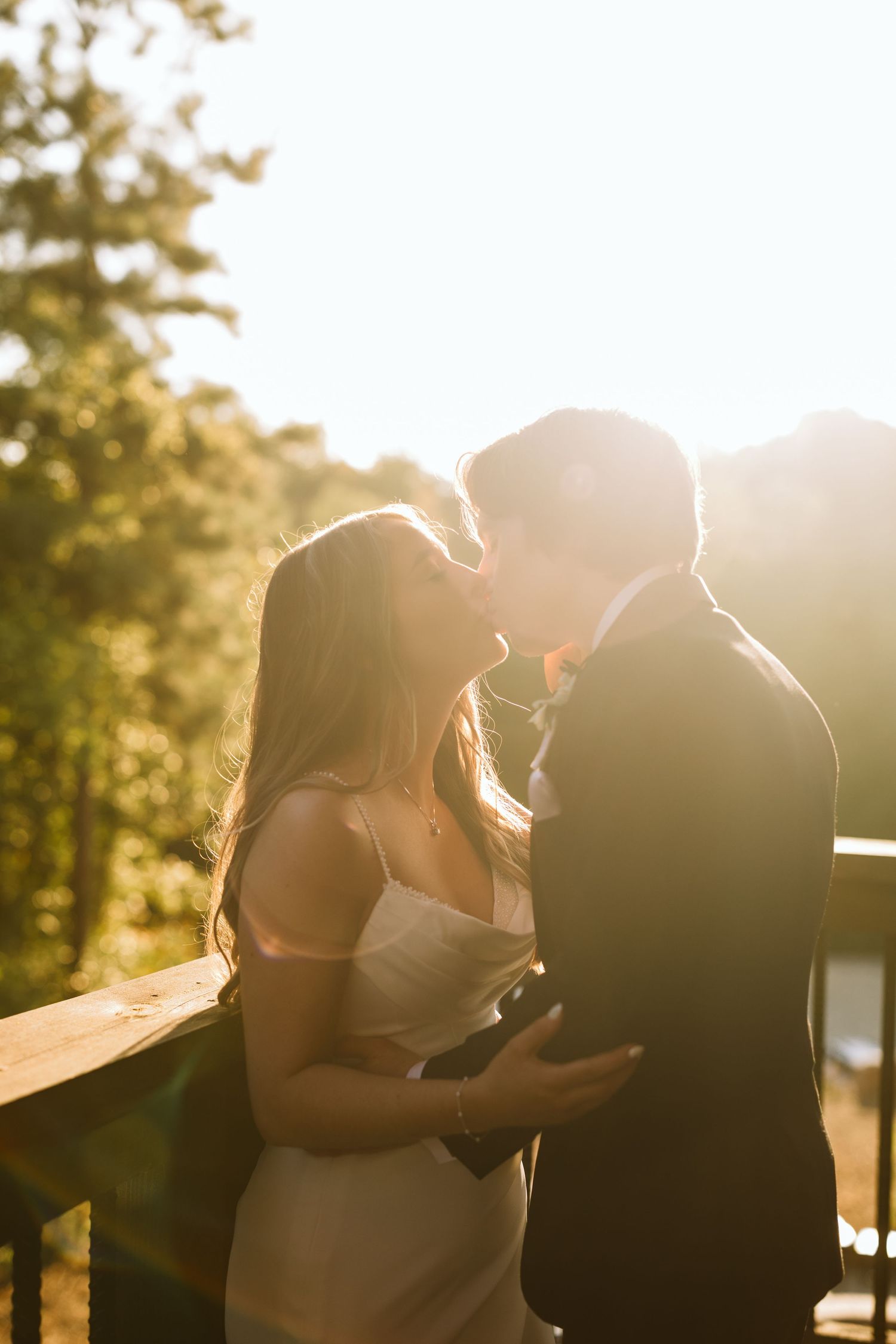 Bride and Groom share a kiss near the balcony railing of the woods with a sun-flare