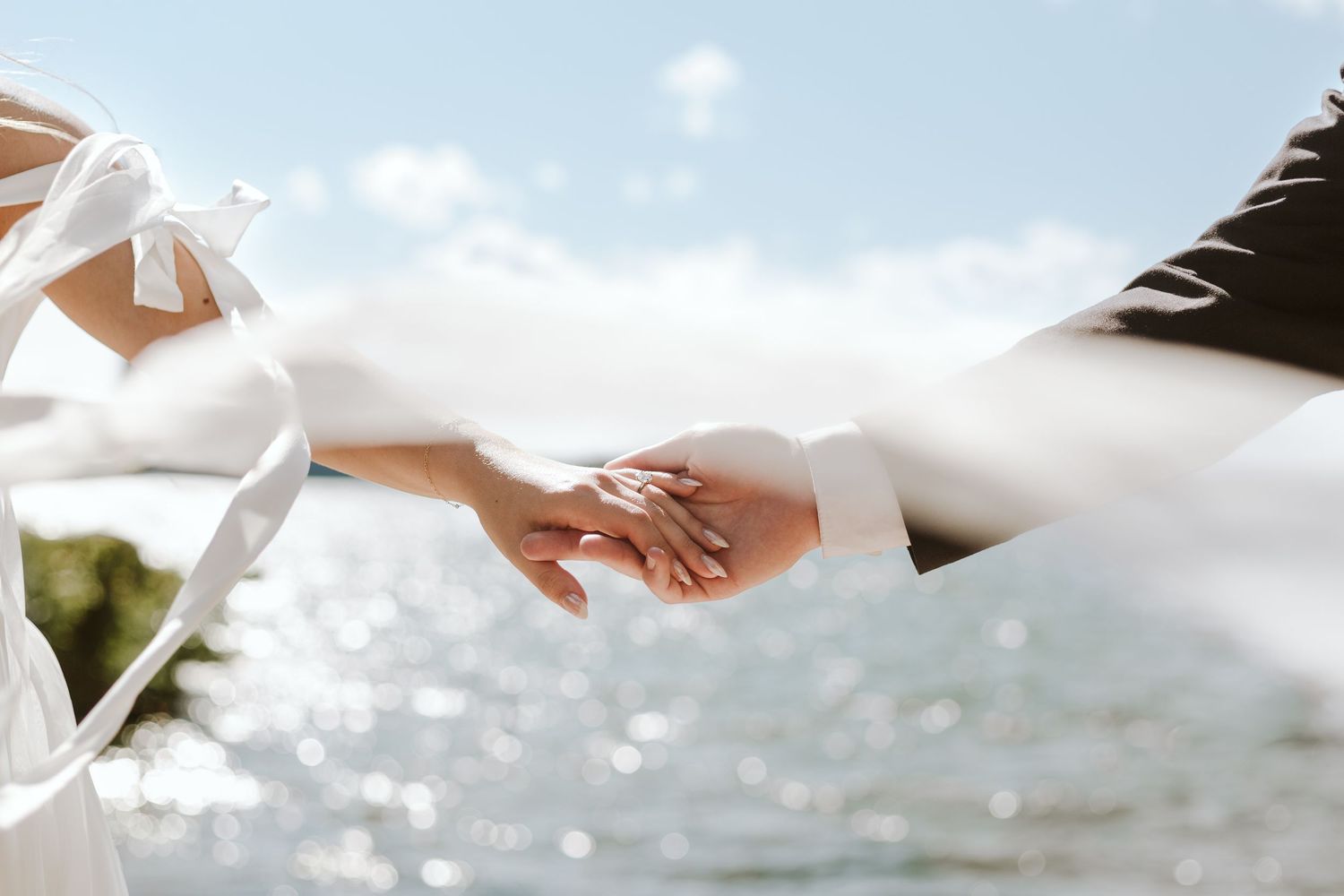 A set of hands holding. One with a suit and white shirt, the other with flowing white straps on her dress with the sun reflecting off water