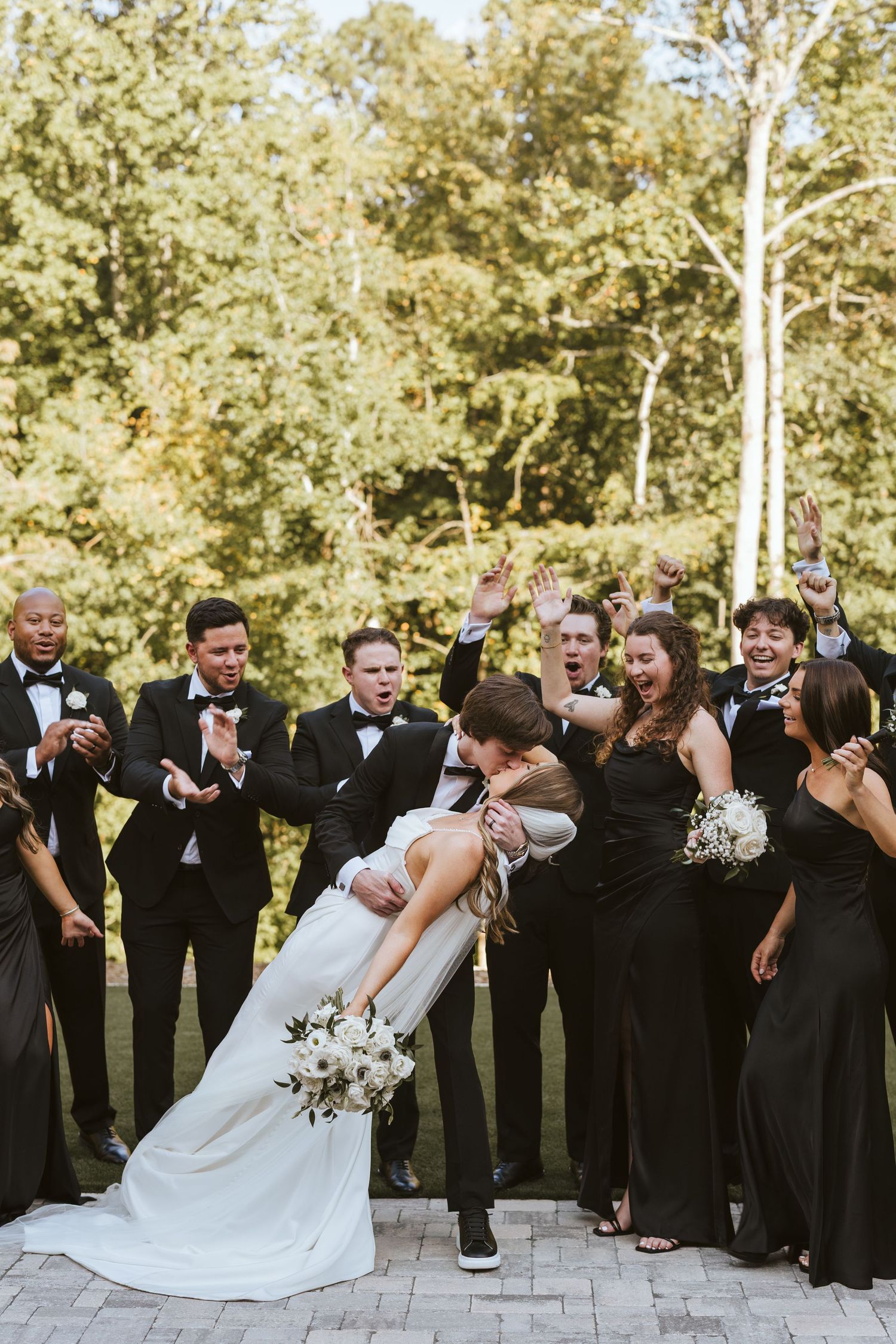Groom dipping his Bride sharing a kiss in front of their cheering wedding party near the tree line