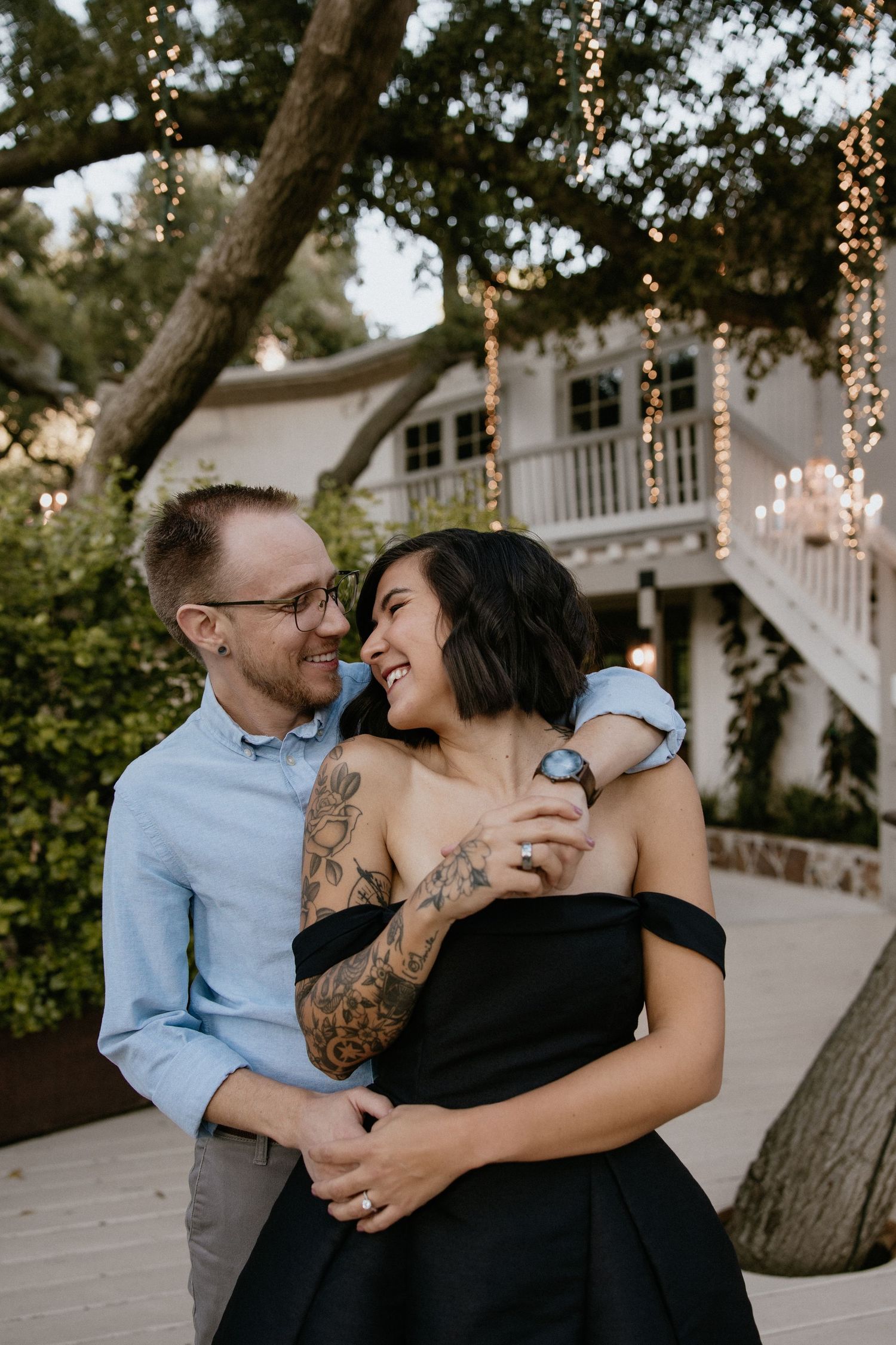 Engaged couple holding each other smiling into each other's eyes in front of a tree and cottage wrapped in lights