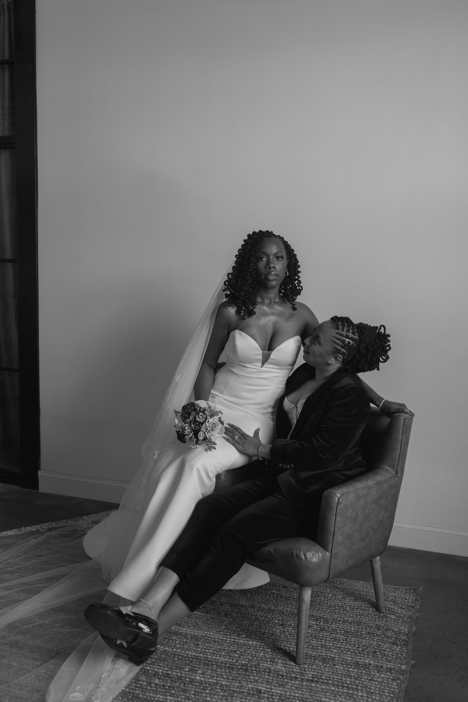 Bride in sleek wedding dress sitting in the lap of her wife in a modern-arm chair