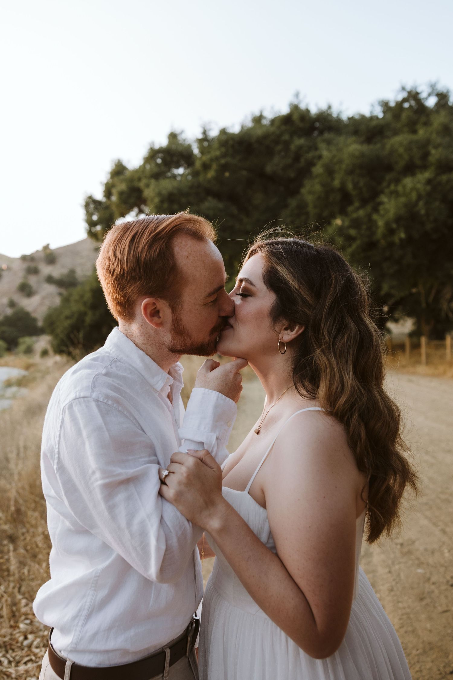red-head man with beard in a white button up shirt embracing his fiancee in white dress while in a field near a tree line