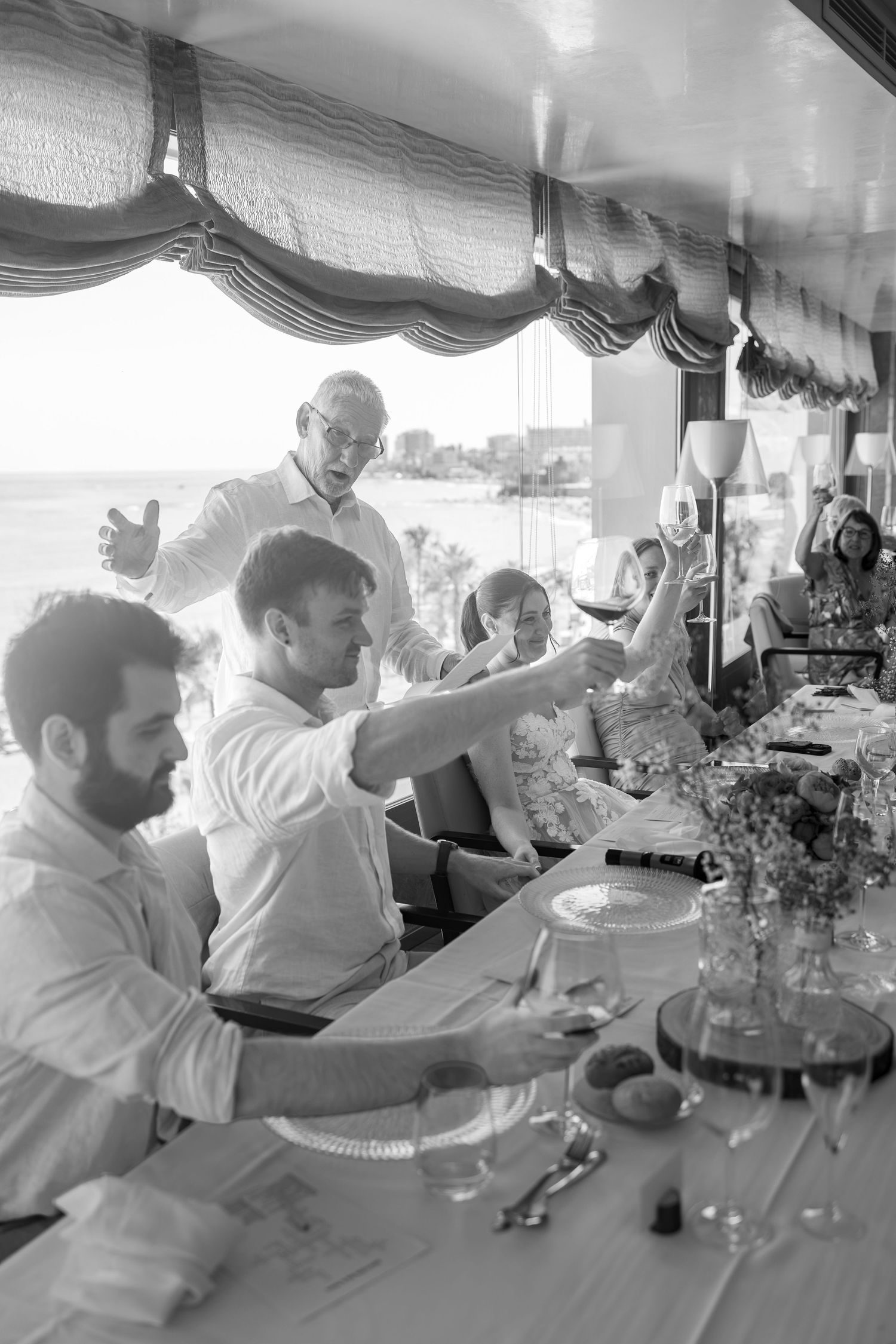 Black and white photo of wedding guests toasting with wine glasses at an elegant seaside restaurant.