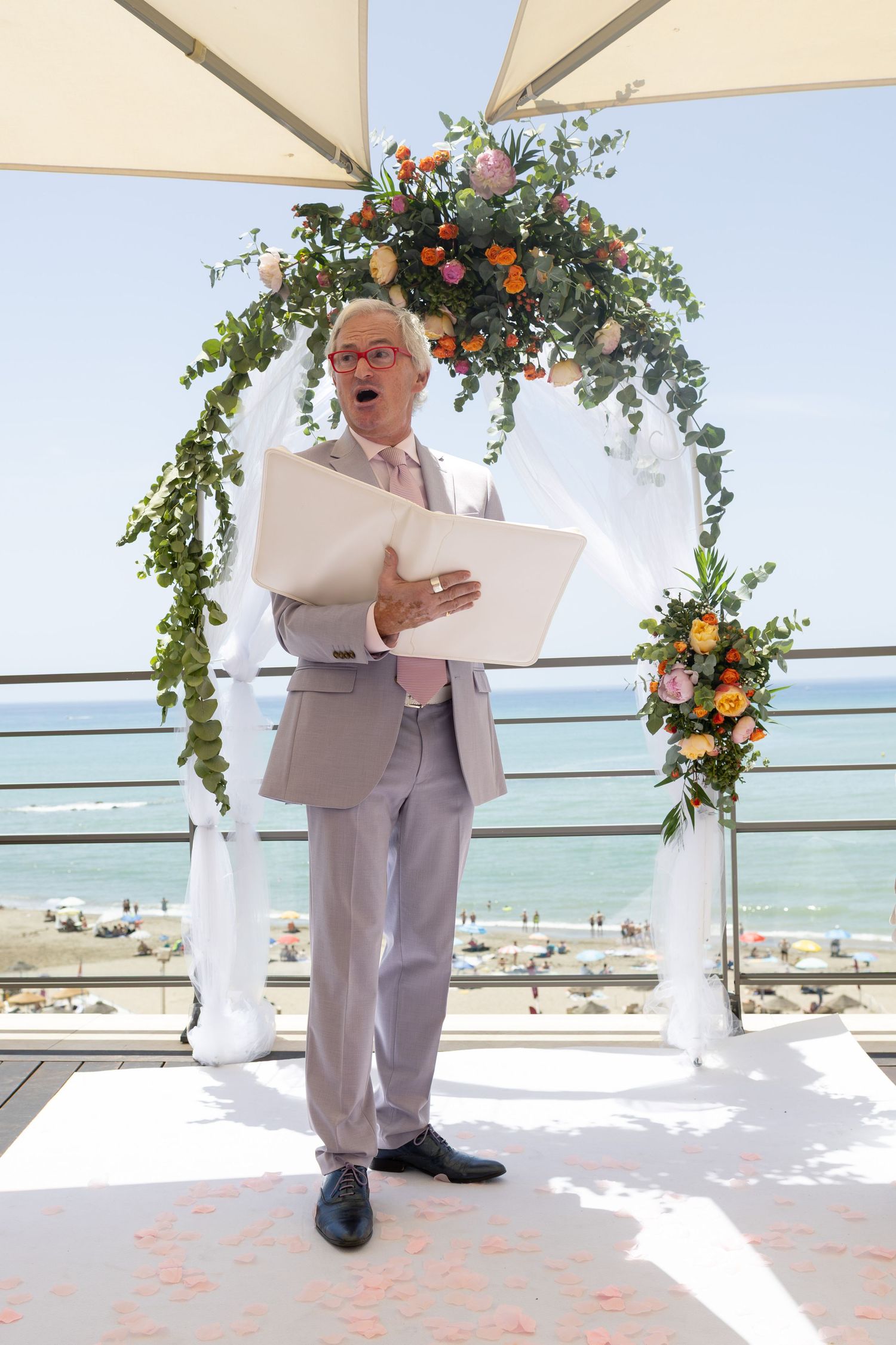 Officiant in grey suit reading from folder at outdoor beach wedding ceremony under floral arch.