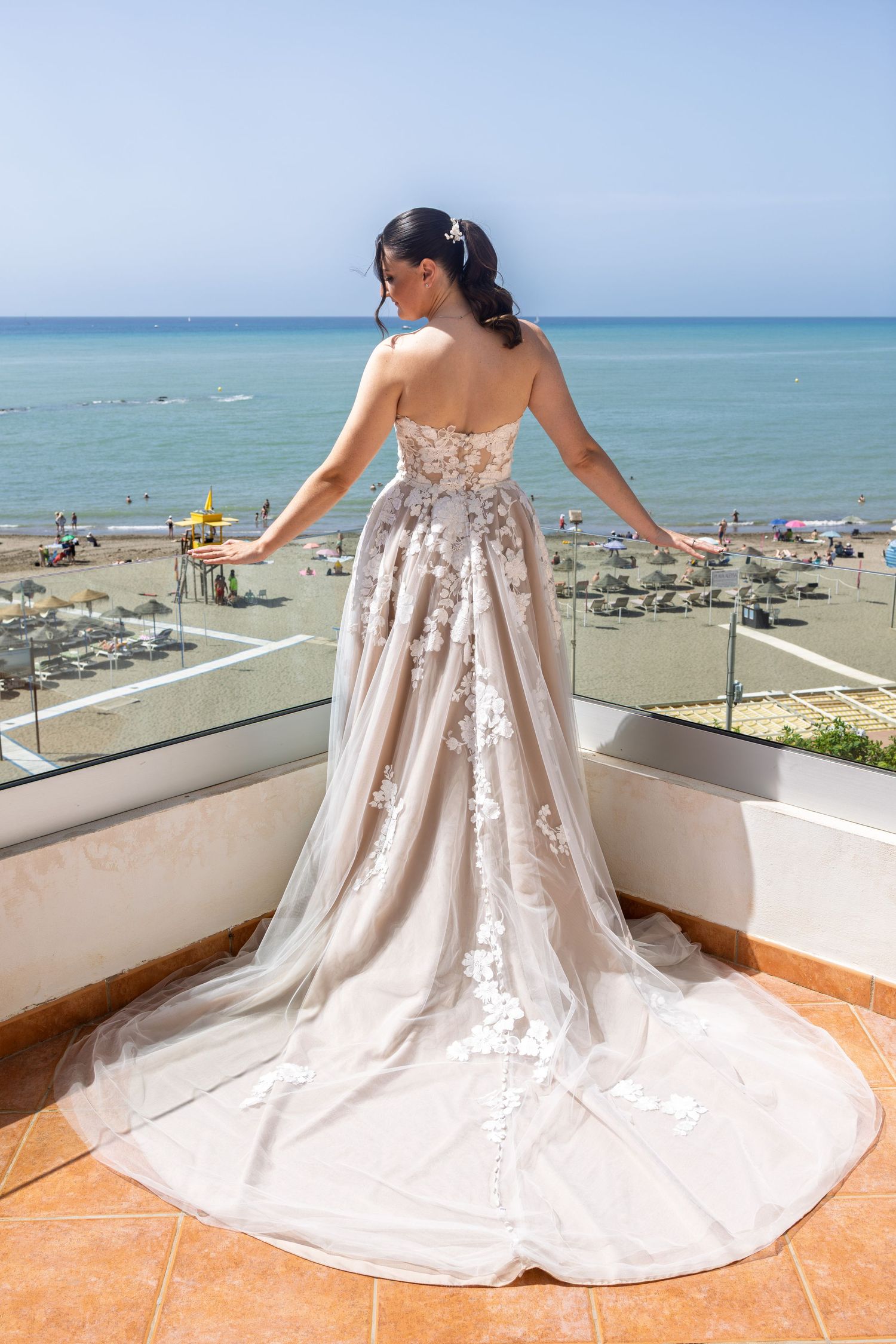 Bride wearing an elegant floral applique wedding dress poses on a terrace with ocean and beach views behind her.