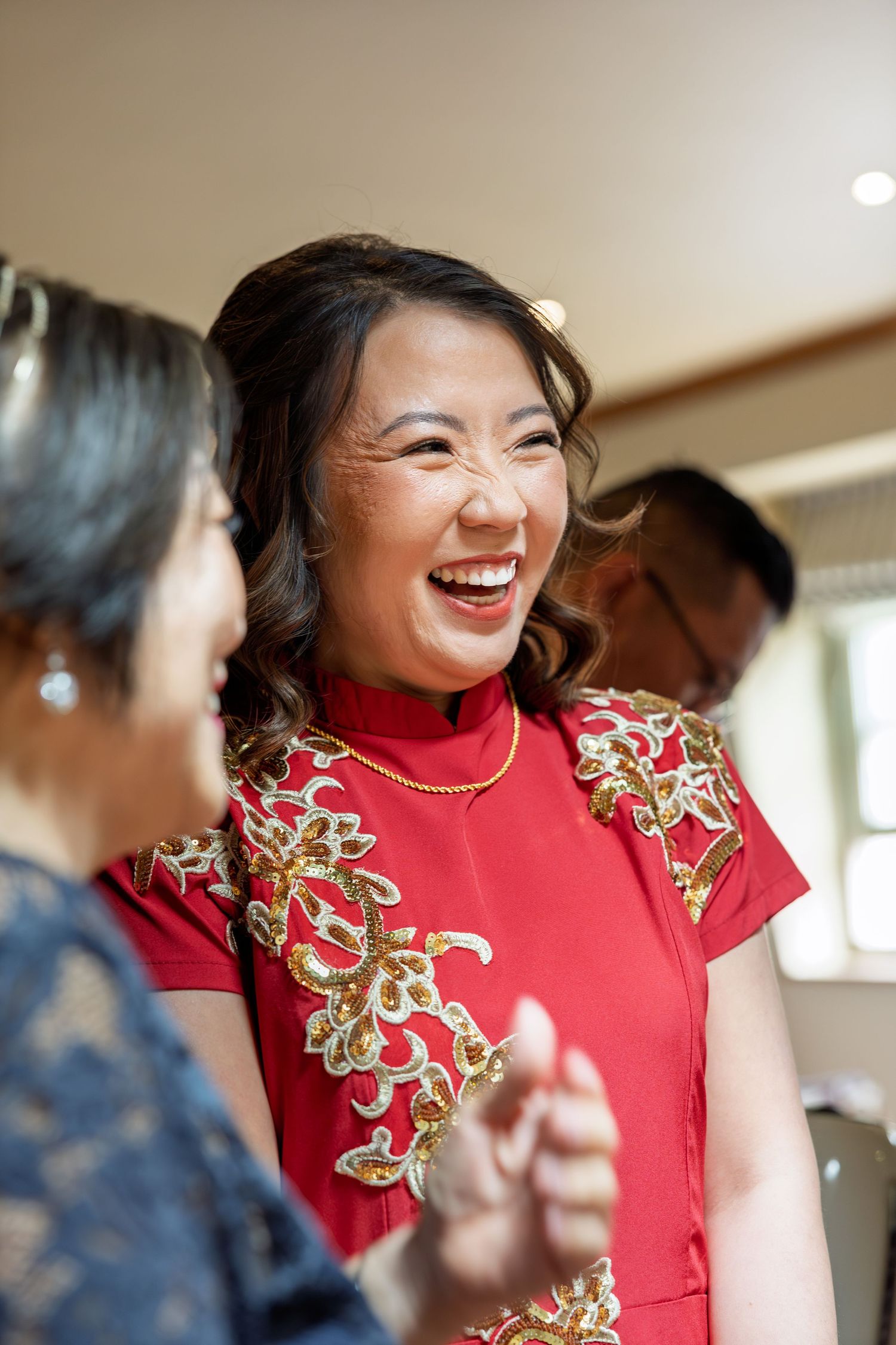 Joyful bride laughing in a red Chinese qipao dress with gold floral embroidery during wedding celebrations.