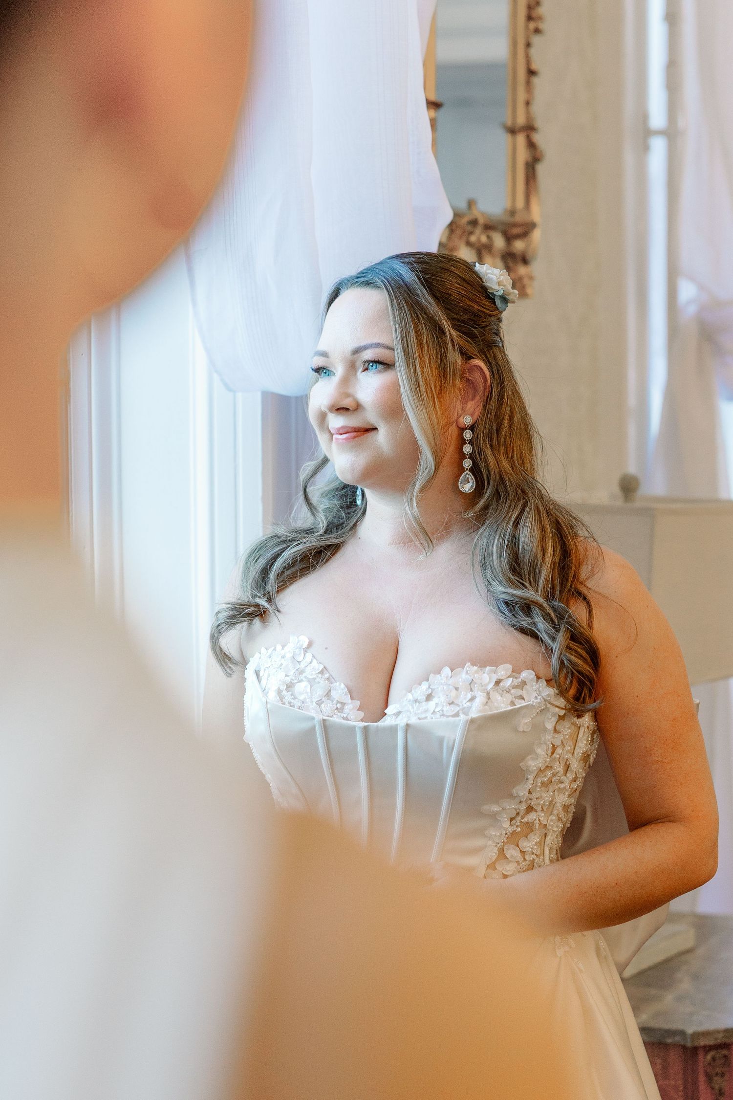 Bride in a white lace wedding dress poses elegantly by a window in a bright, classic interior setting.
