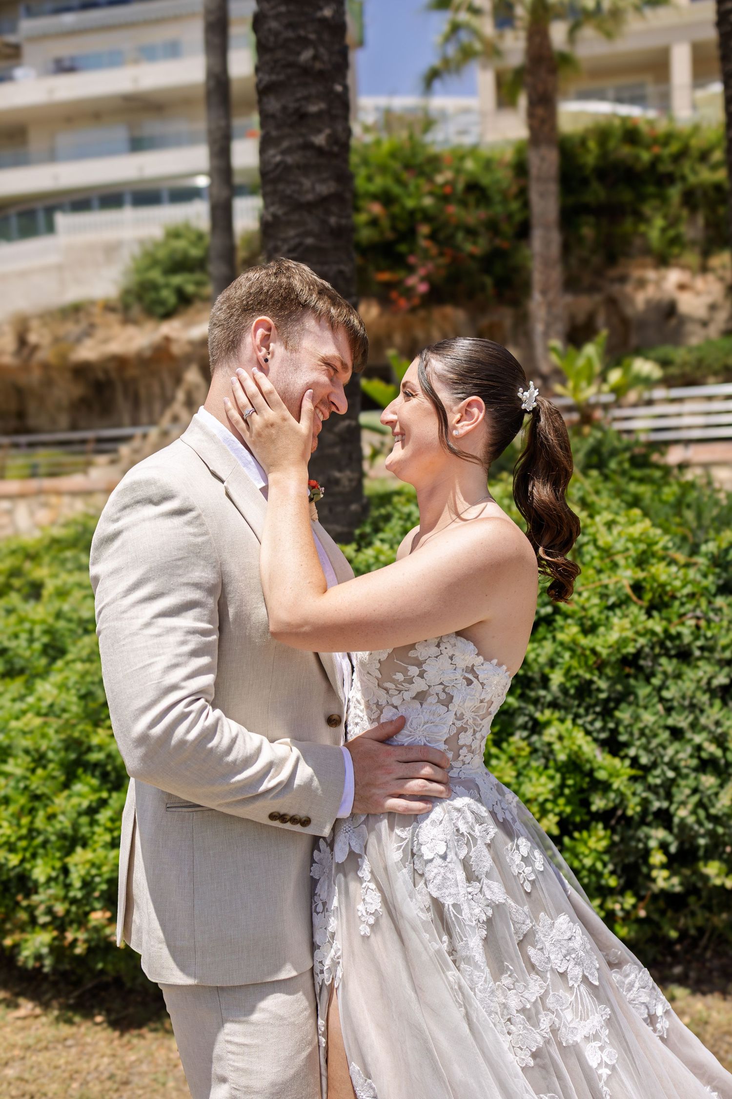 Smiling bride and groom share a tender moment outdoors, with the bride touching the groom's face in a lush garden setting.