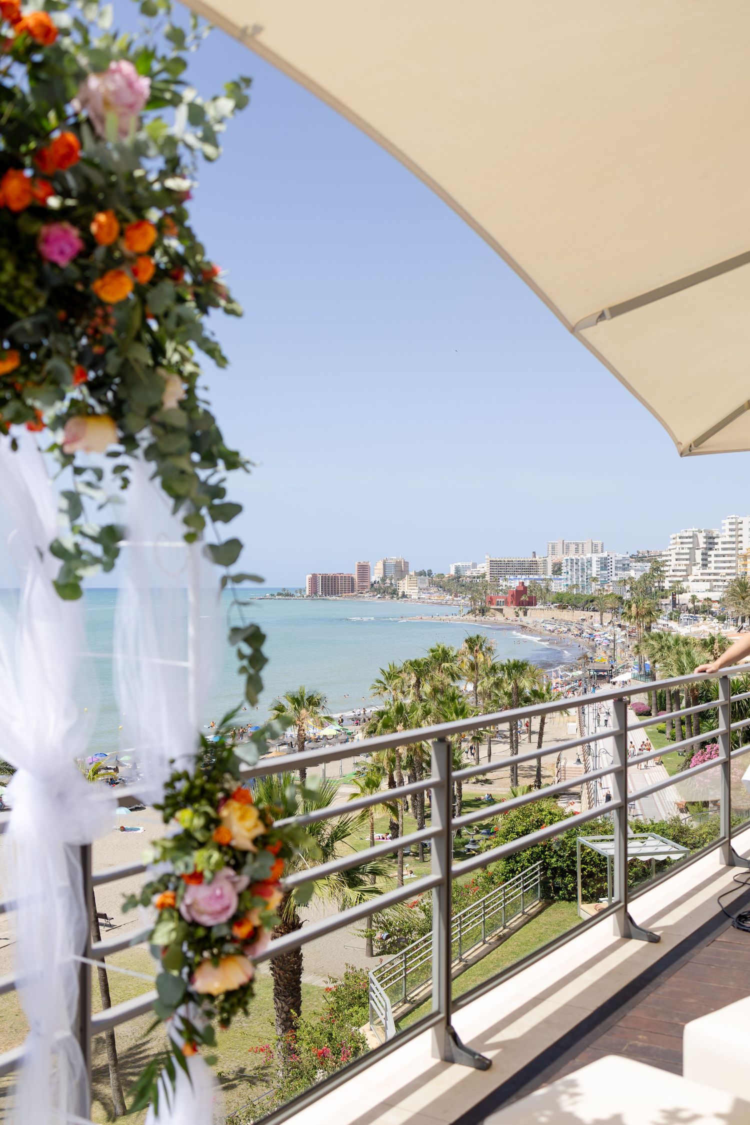 Colorful floral wedding arch frames a stunning coastal view of a sunny beach and turquoise Mediterranean sea.