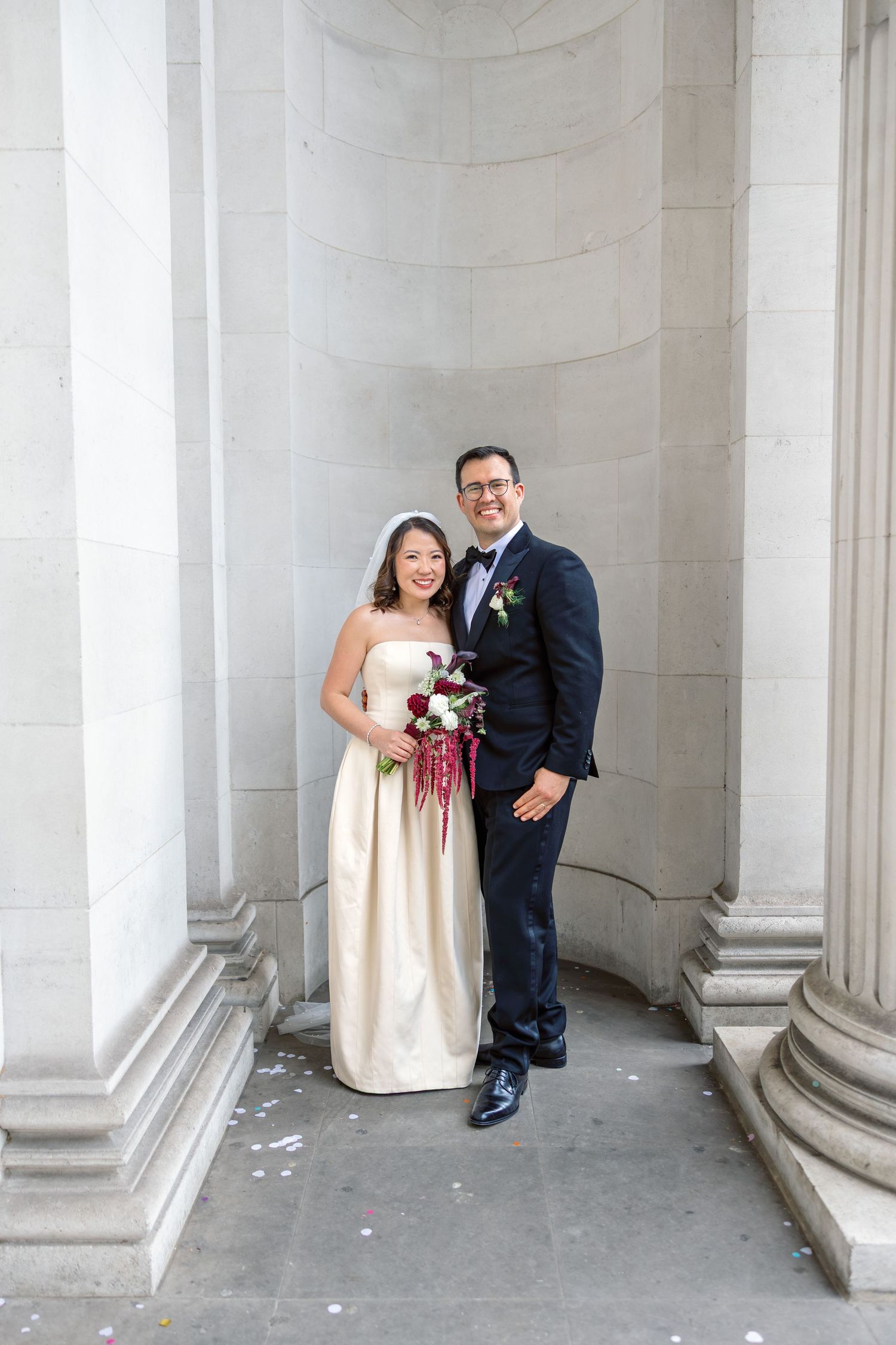 Bride in strapless gown and groom in navy suit pose with bouquet between stone columns at elegant wedding venue.