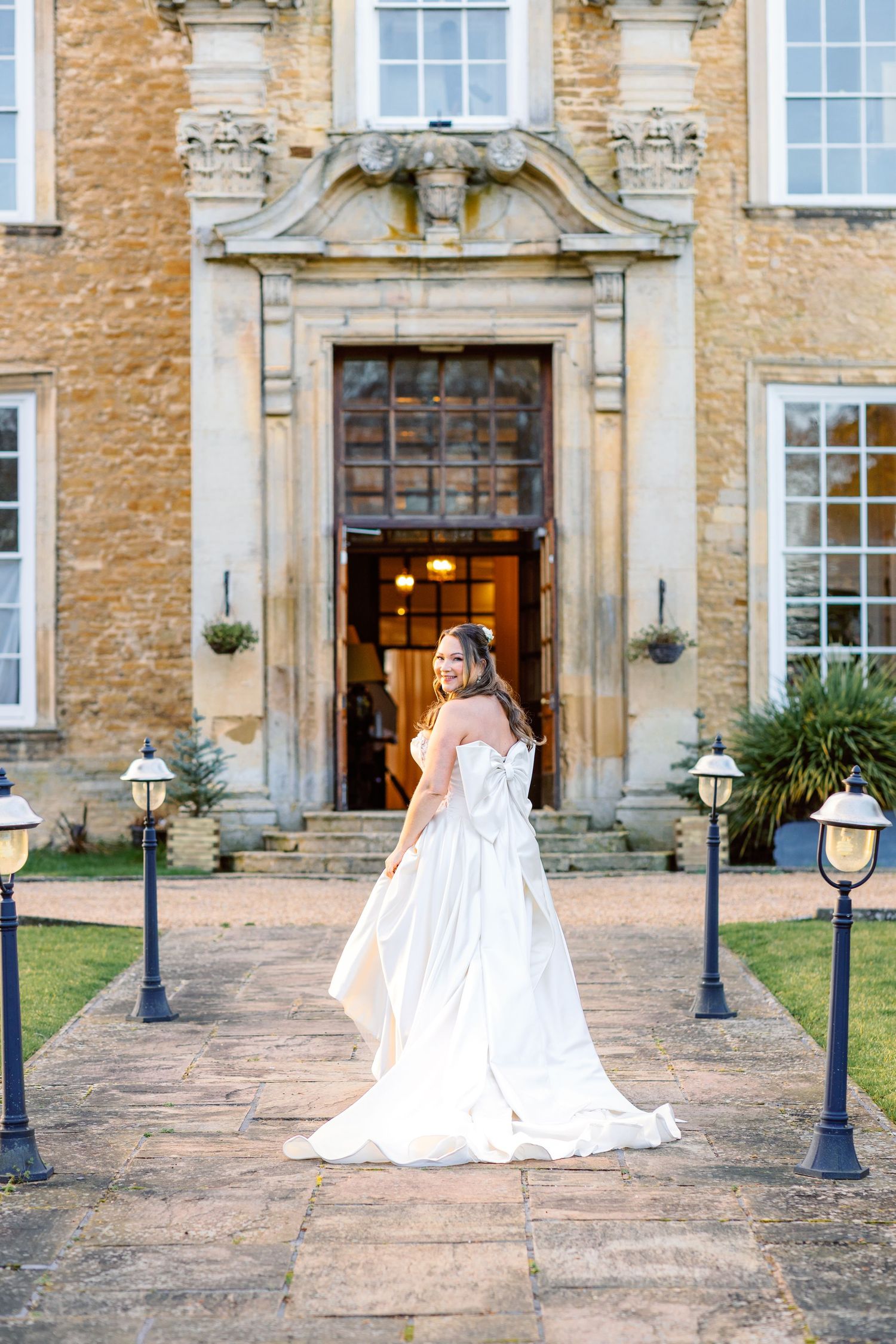Bride in flowing white gown standing before grand stone mansion entrance with ornate architecture.