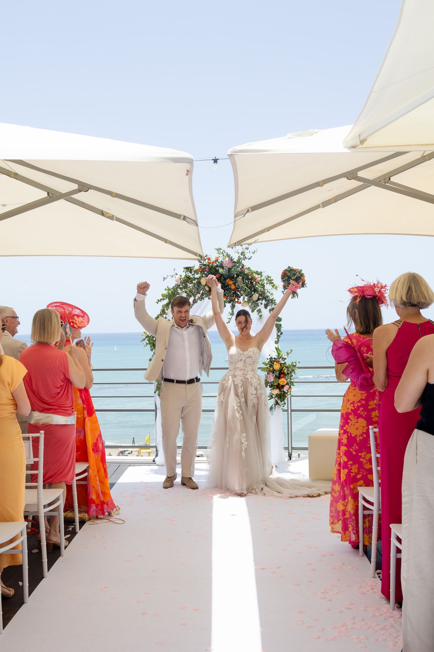 Joyful newlyweds raise arms in celebration at outdoor beachside wedding ceremony with colorful guests seated along aisle.