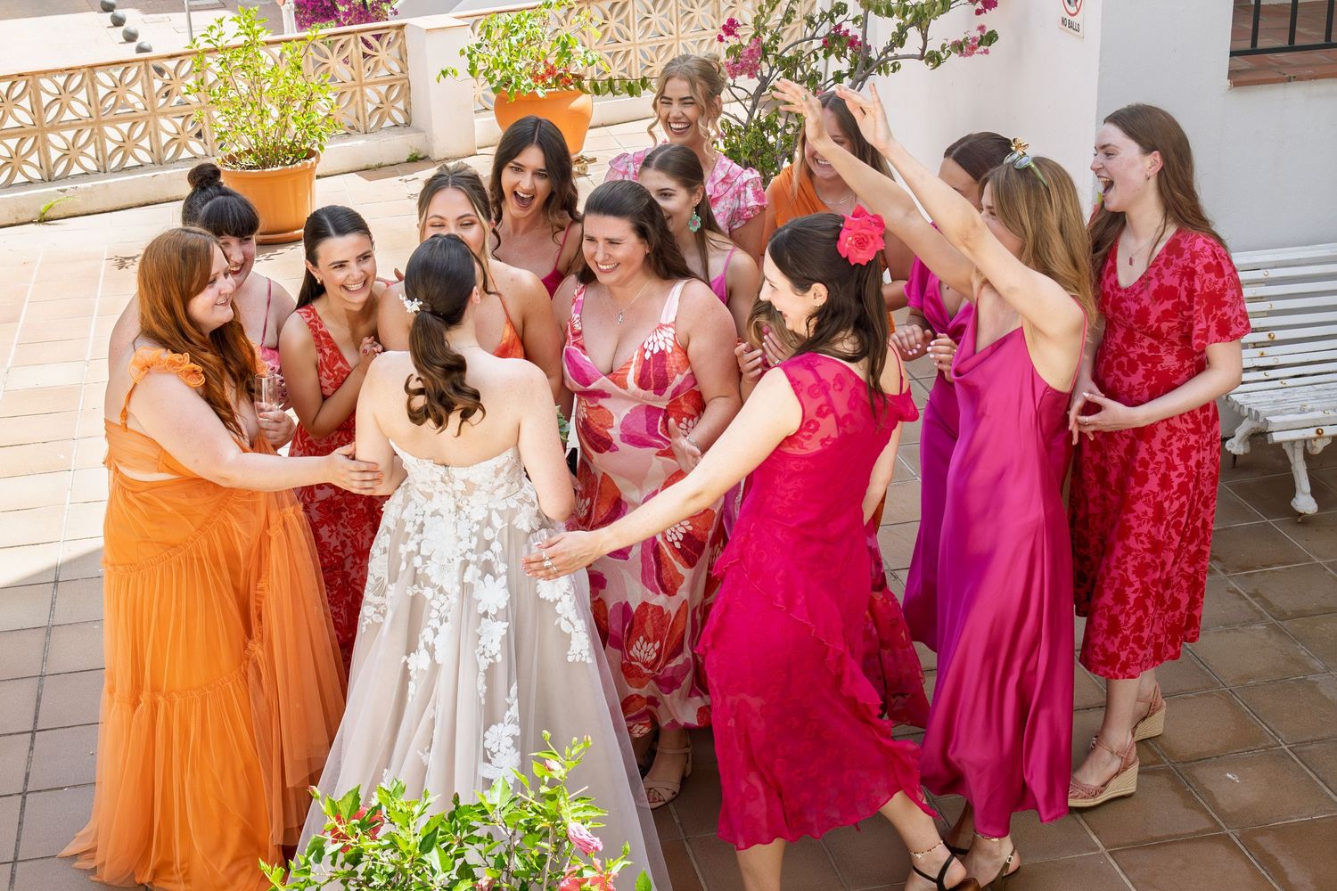 Bride surrounded by bridesmaids in colorful red and pink dresses celebrating at an outdoor wedding venue.