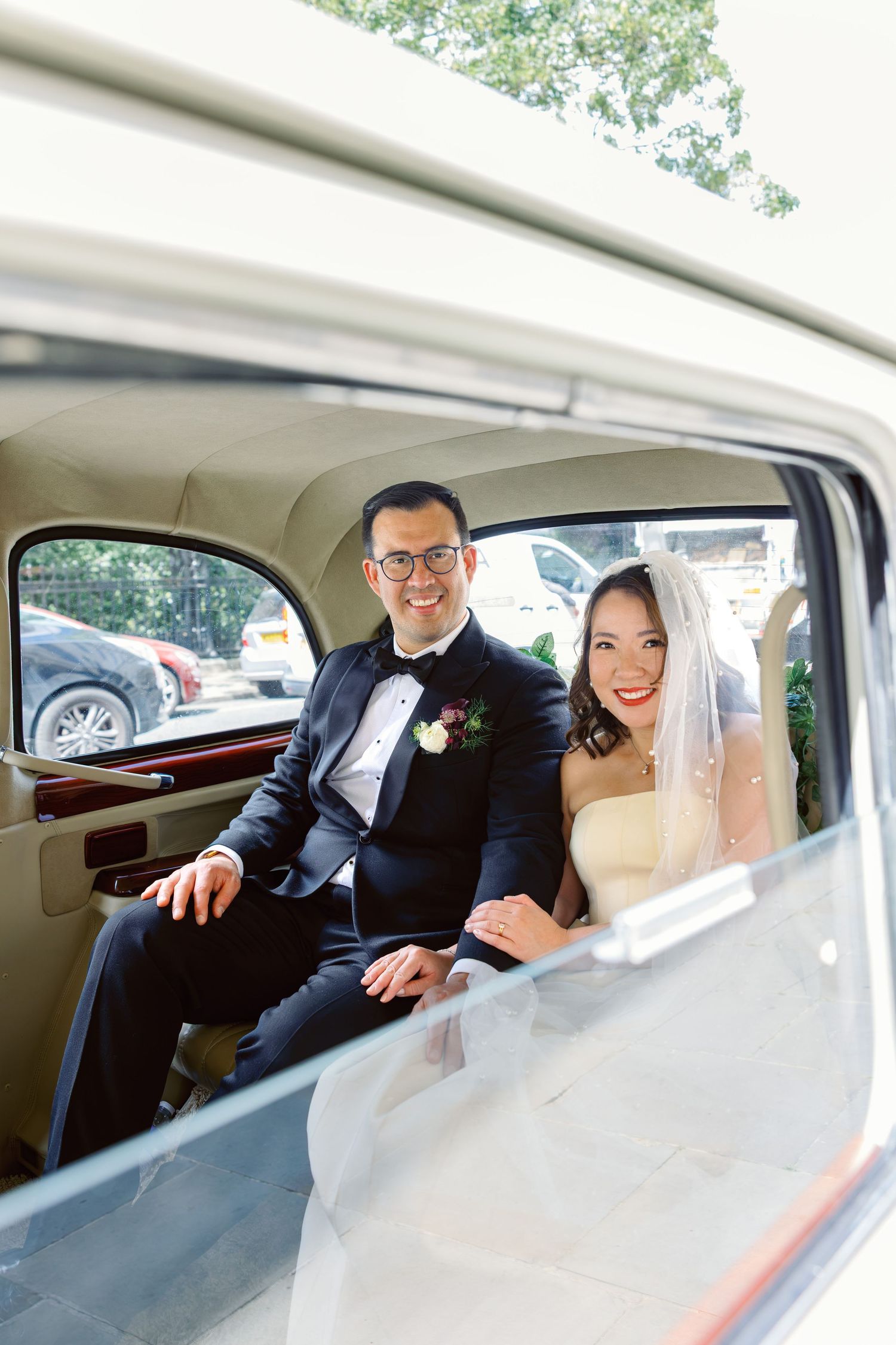 Smiling bride and groom seated inside a classic vintage wedding car viewed through the window.