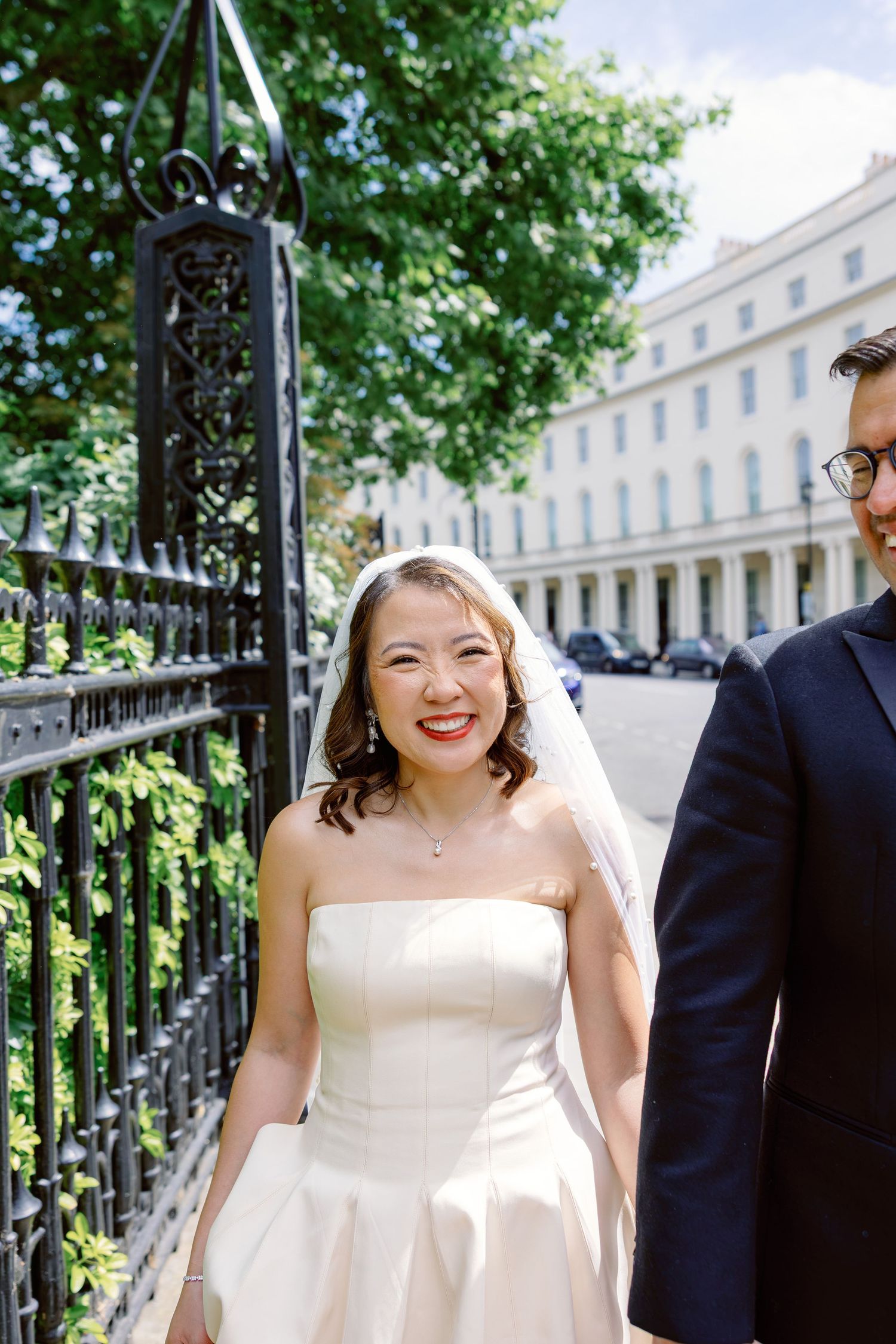 Radiant Asian bride in strapless white wedding dress smiling joyfully beside iron gates on wedding day.