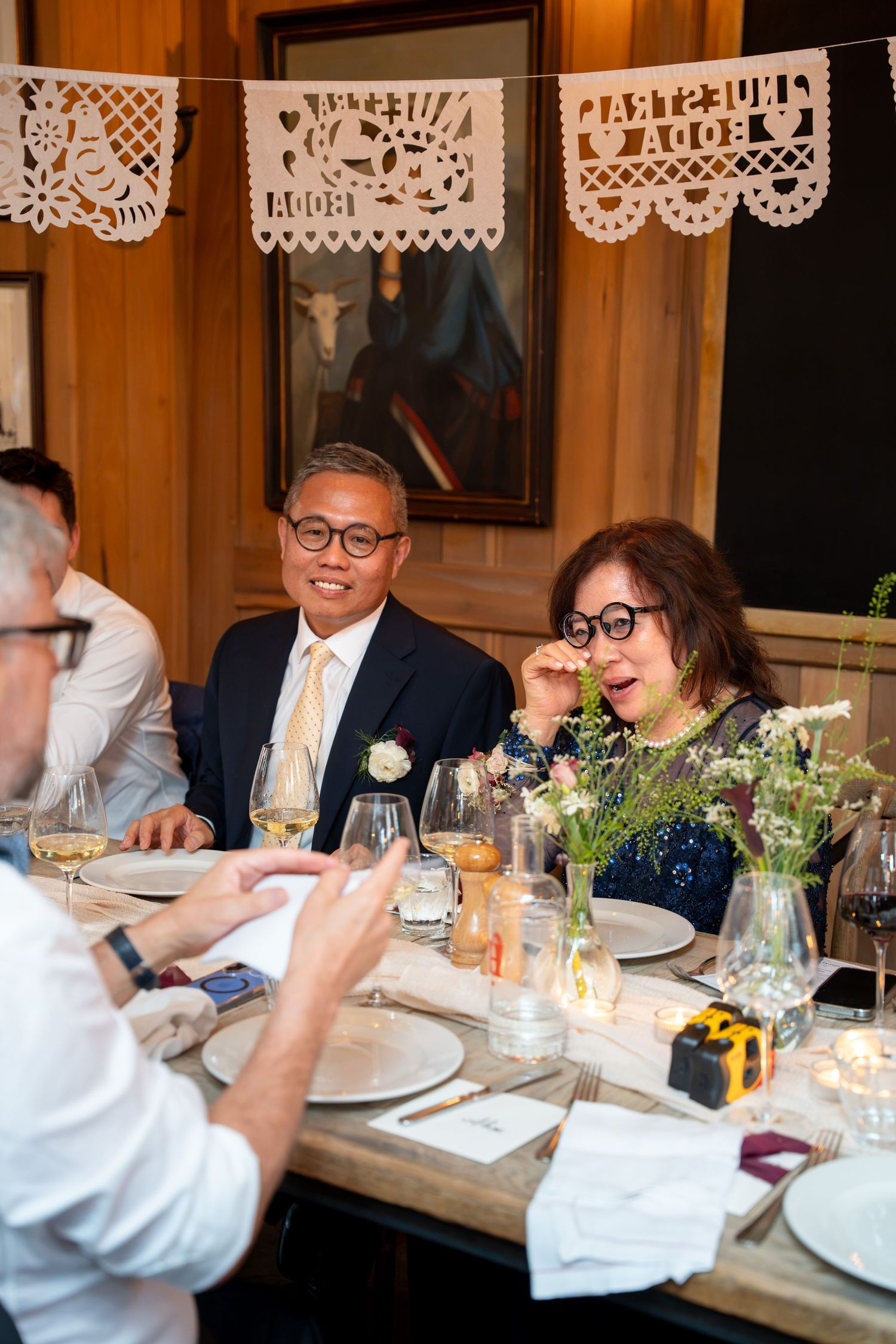 Wedding guests laugh and converse at elegantly decorated reception dinner table with floral centerpieces.