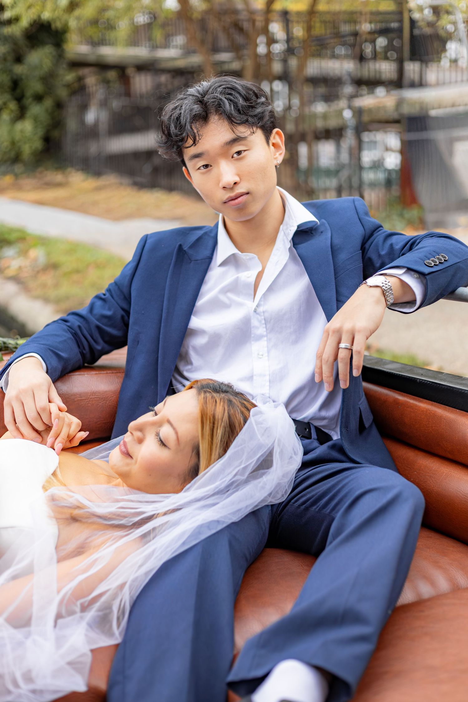 Couple in wedding attire poses romantically on brown leather seating during outdoor photo session.