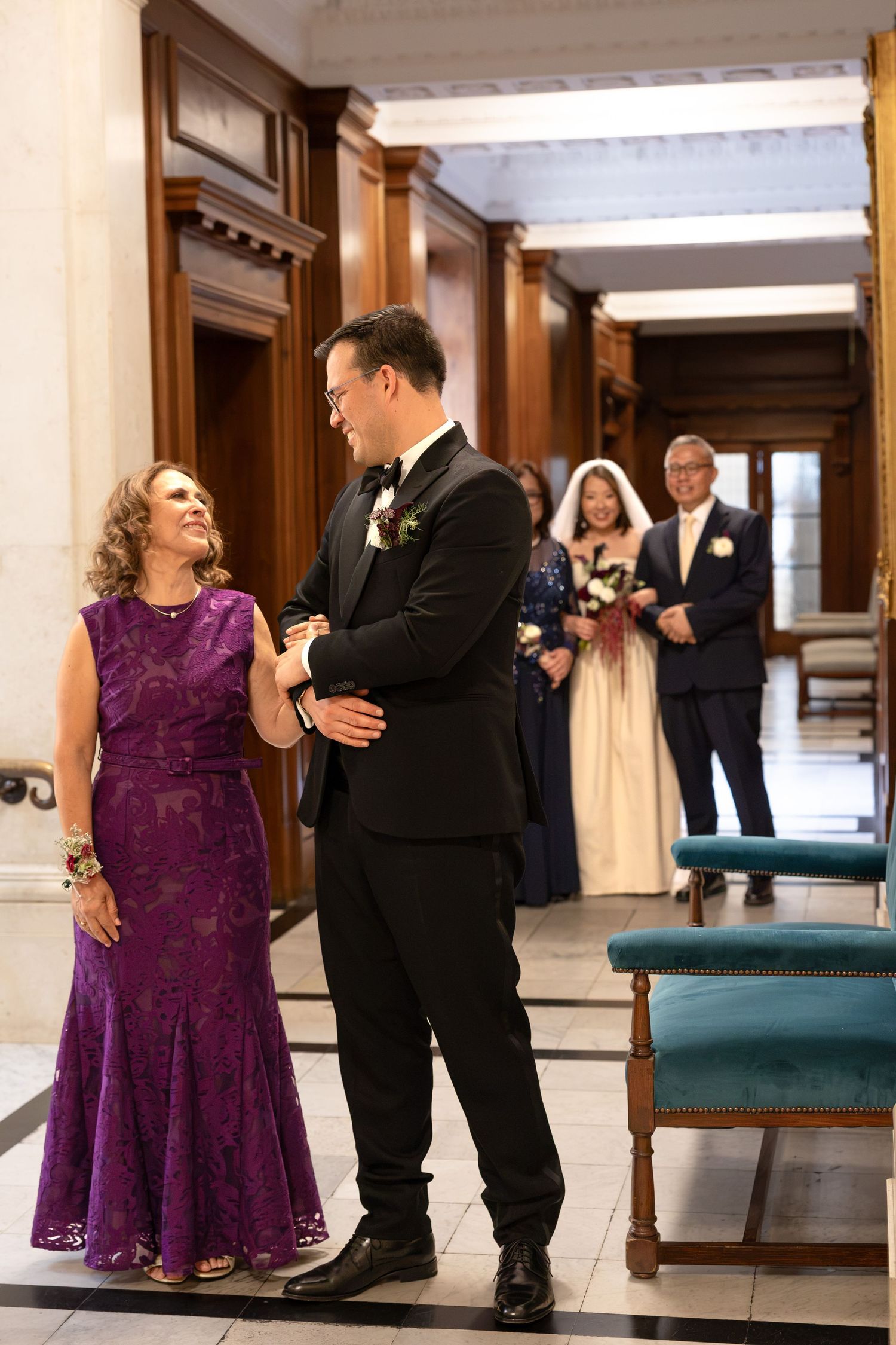 Groom in tuxedo embraces mother in purple dress while bride approaches with parents in background.