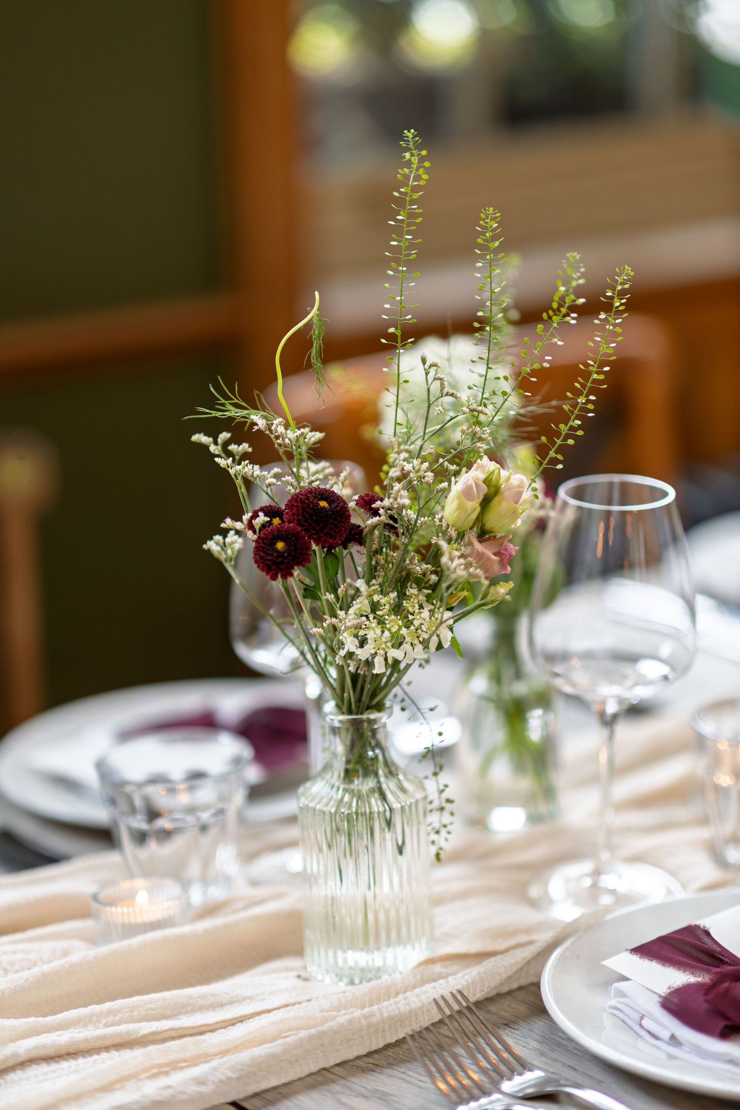 Elegant wedding table centerpiece with burgundy and pink wildflowers in glass vase on cream runner.
