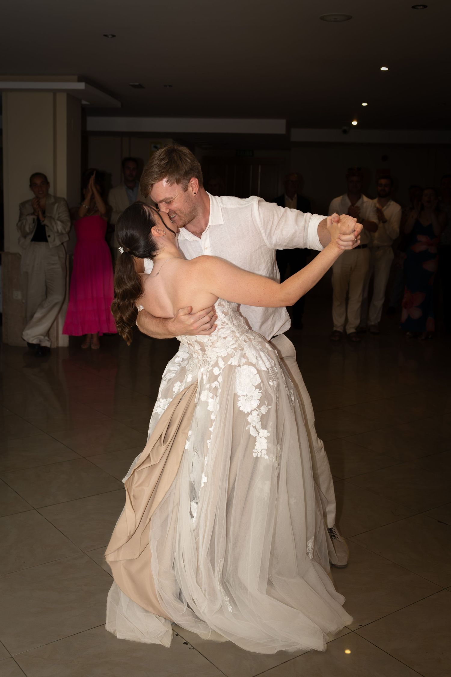 Bride and groom share their first dance, elegantly embracing on the dance floor surrounded by wedding guests.