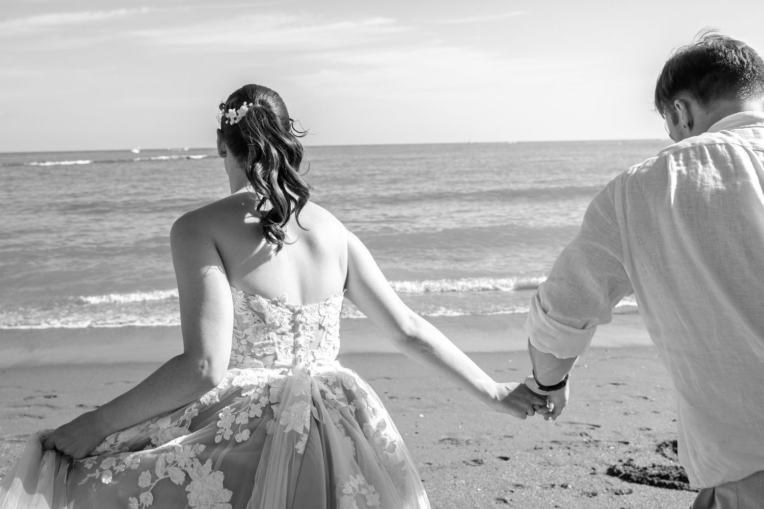 Black and white photo of bride and groom holding hands walking along a sandy beach shoreline.