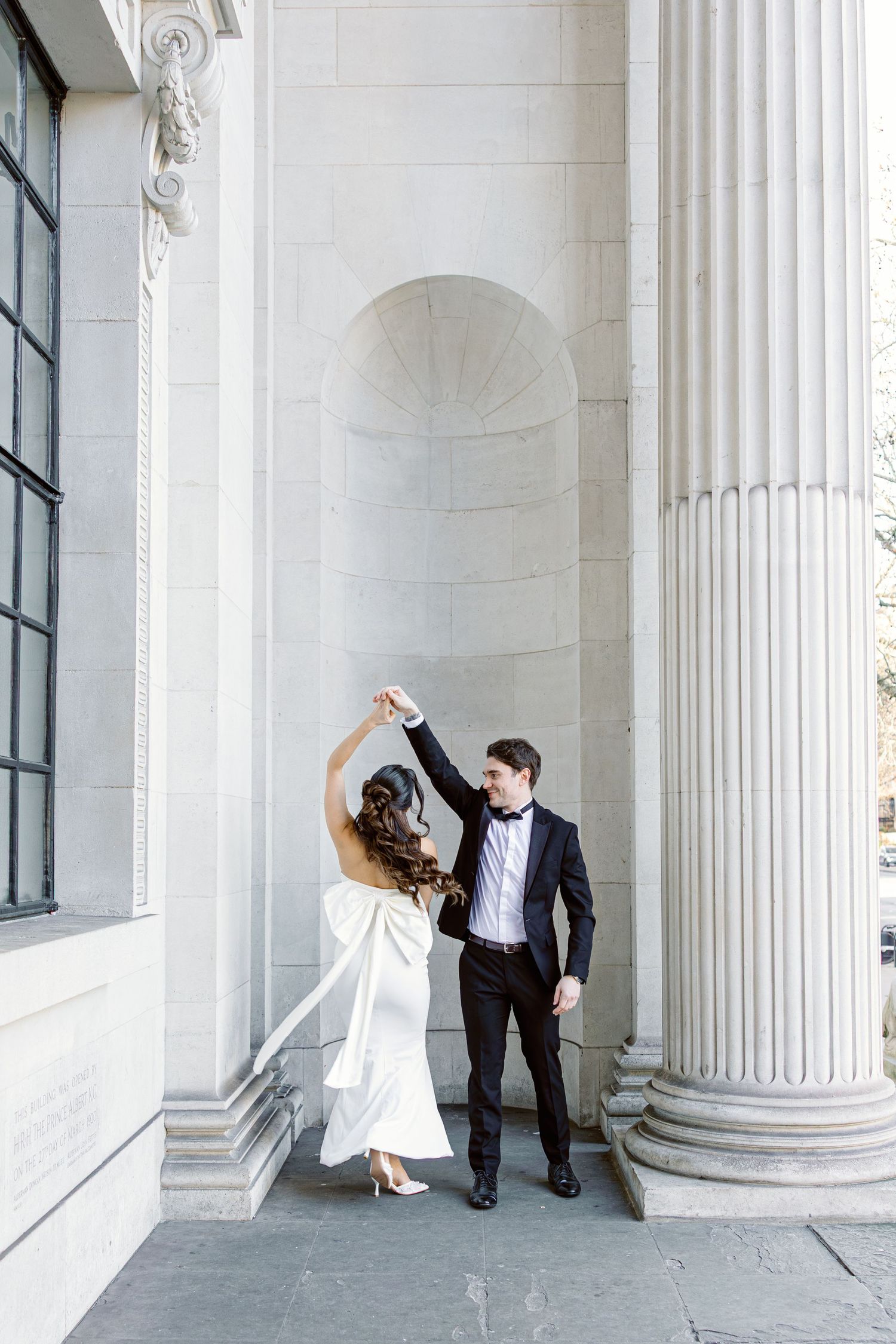 Elegant couple dances playfully between white classical columns in a romantic architectural setting.