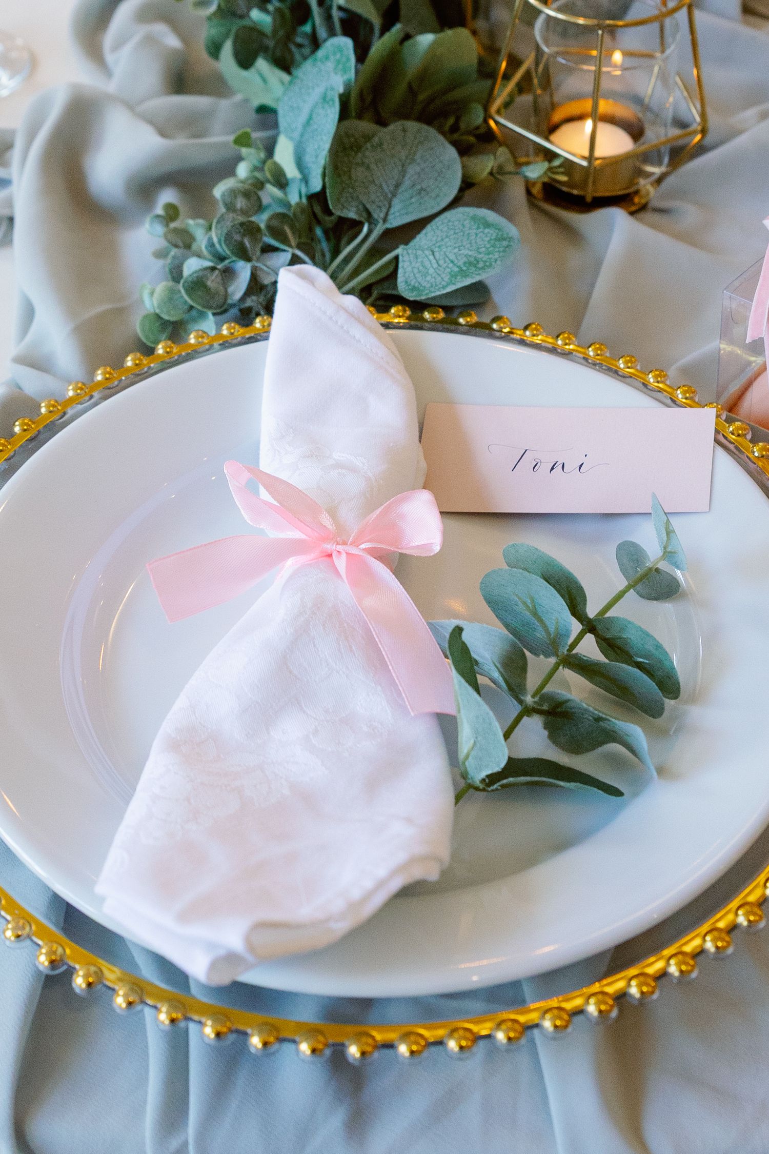 Elegant table setting with gold-rimmed plates, pink napkins tied with bows, and fresh eucalyptus sprigs as decoration.