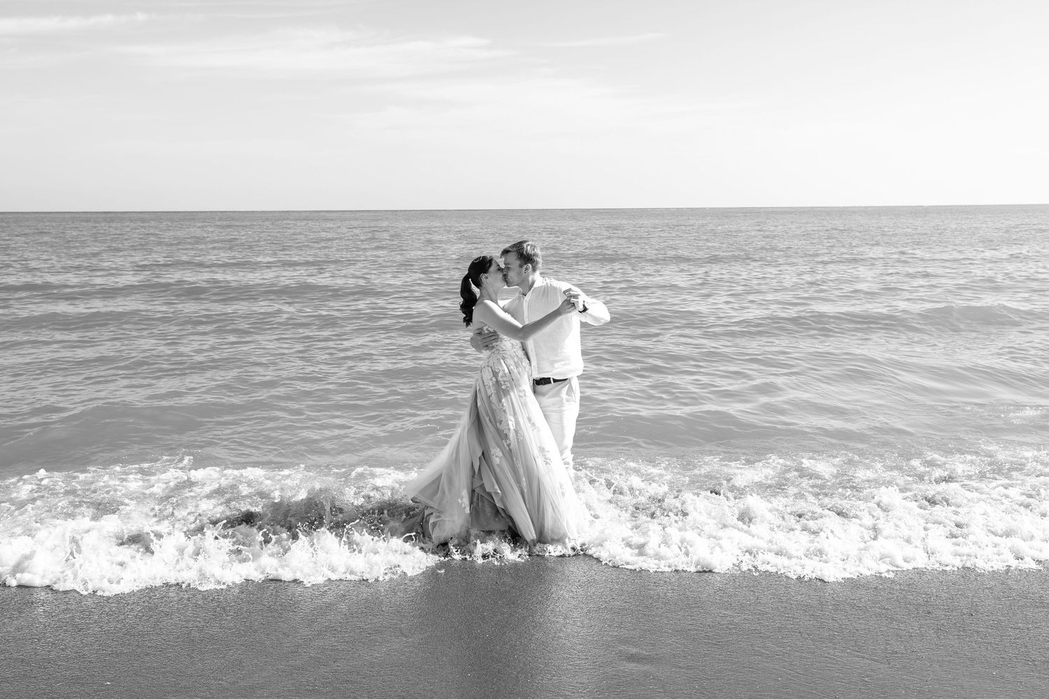 Black and white romantic photo of bride and groom embracing in ocean waves on their wedding day.