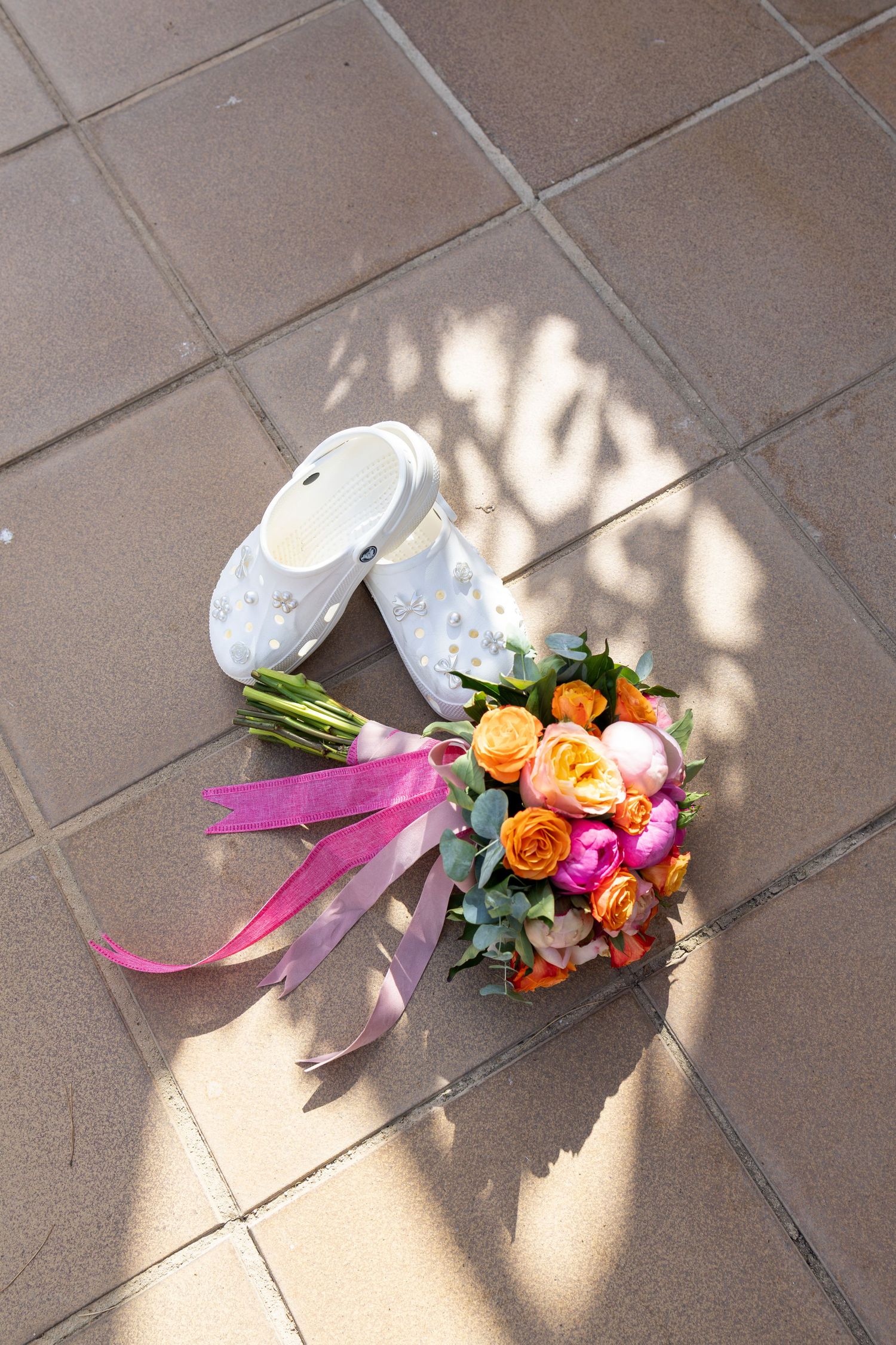 Bridal bouquet of vibrant orange and pink roses with pink ribbons beside white Crocs on tiled ground.