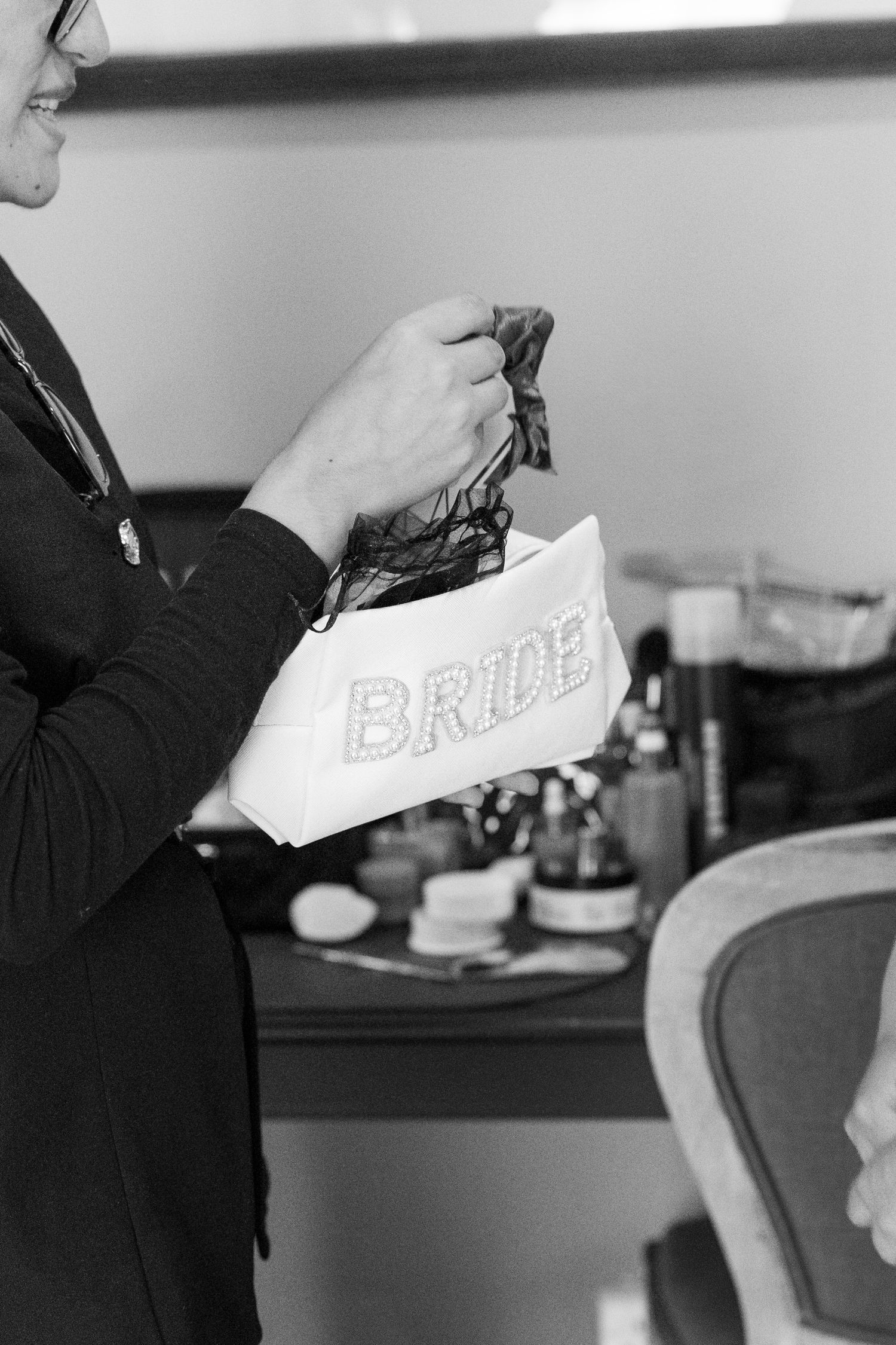 Black and white photo of a bride holding a white cosmetic bag labeled BRIDE at a wedding getting-ready session.