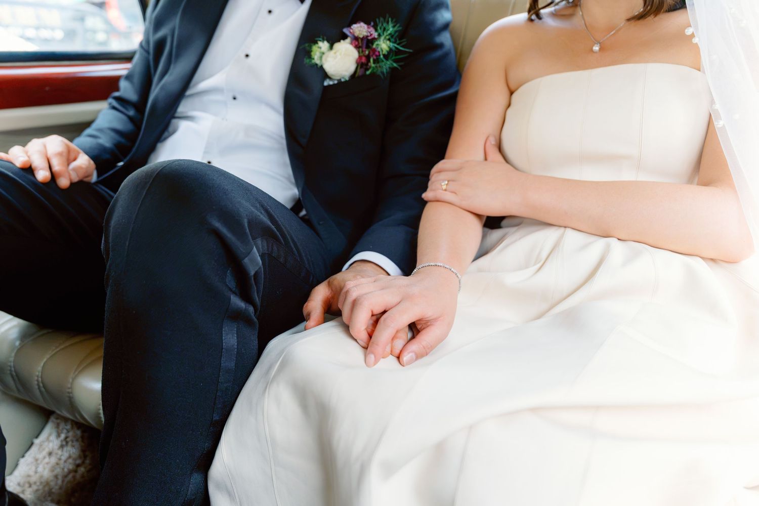 Close-up of bride and groom holding hands, bride in strapless white dress, groom in navy suit with floral boutonniere.