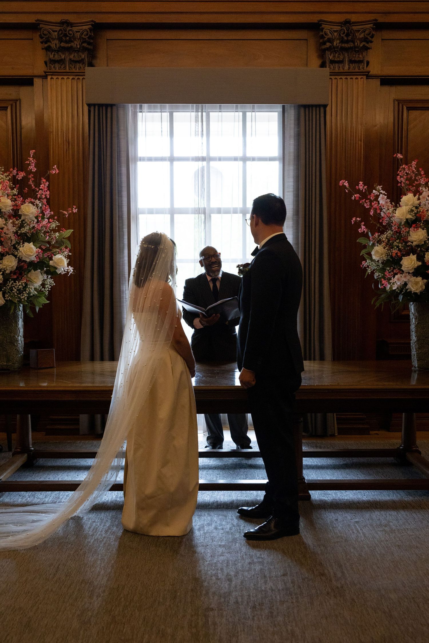 Bride and groom exchange vows at altar flanked by pink floral arrangements in elegant wood-paneled room.