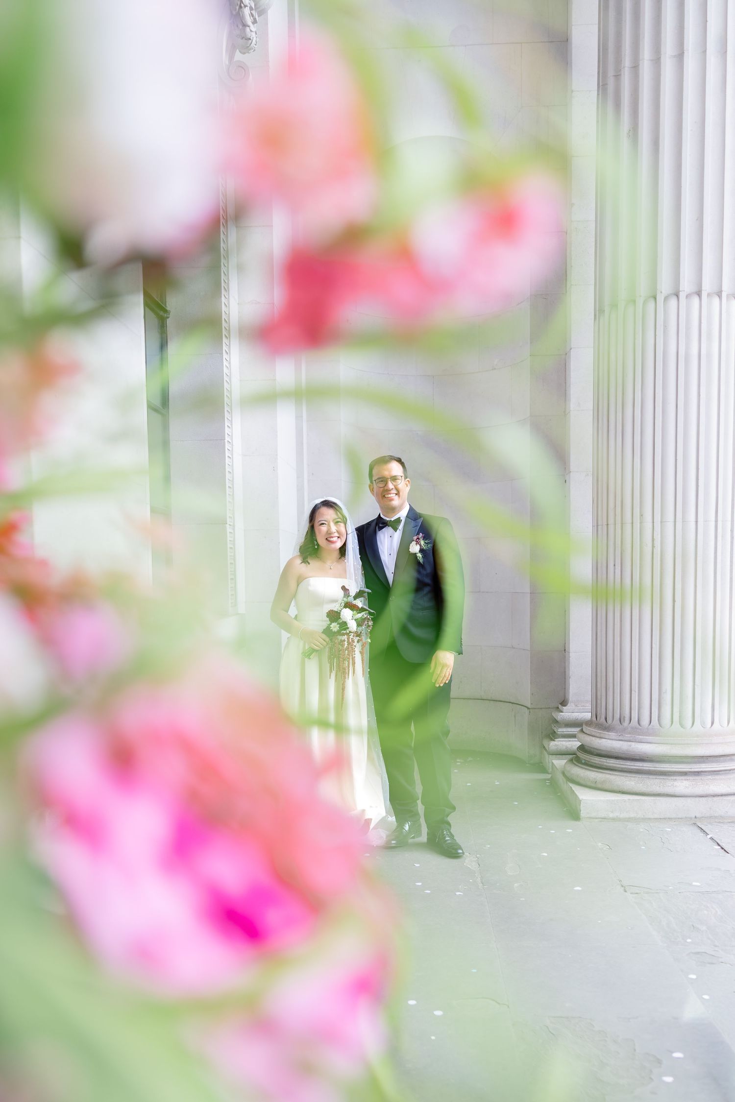 A bride and groom pose between white columns, framed by blurred pink flowers in the foreground.
