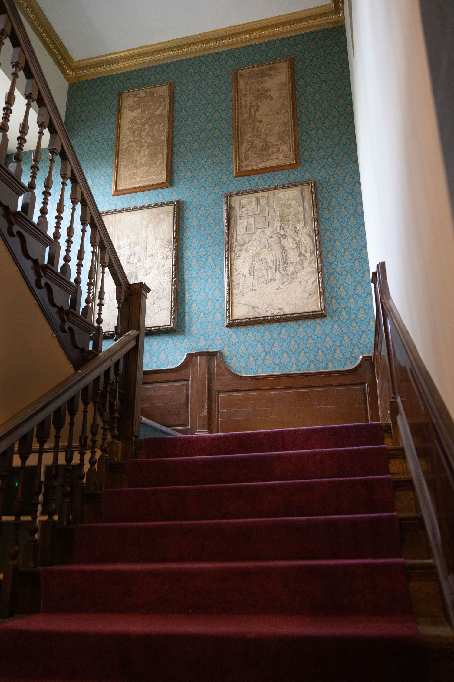 Elegant staircase with red carpet and turquoise wallpaper featuring framed artwork in a historic interior setting.