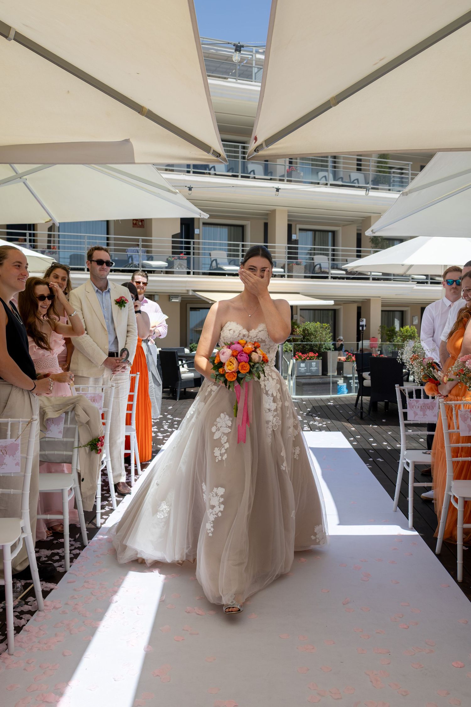Radiant bride walks down outdoor wedding aisle in floral lace gown carrying vibrant orange and pink bouquet.