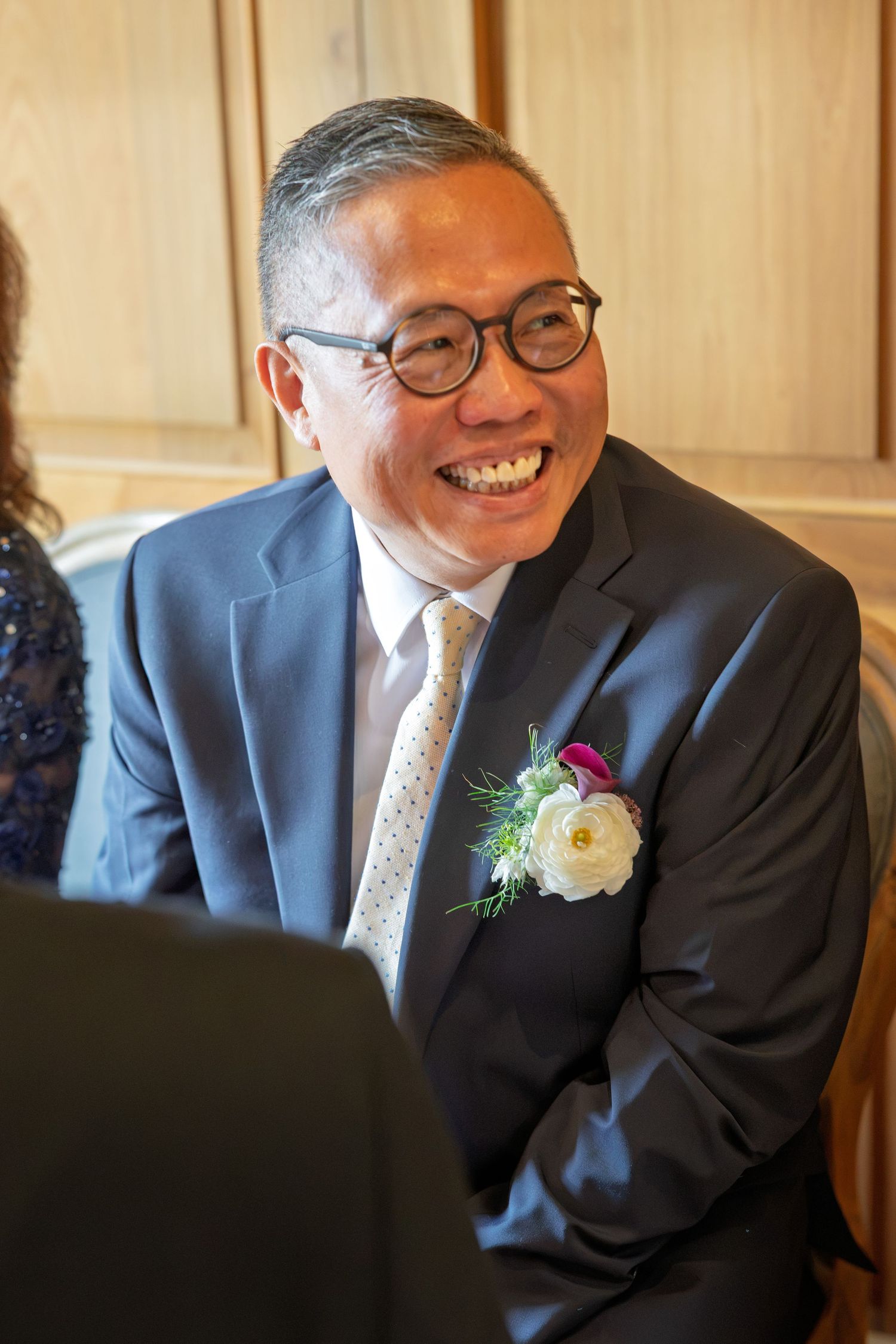 A smiling man in a navy suit with a white floral boutonniere at a wedding celebration.