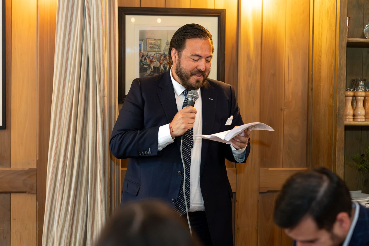 Bearded officiant in navy suit reading from papers into microphone during indoor wedding reception speech.