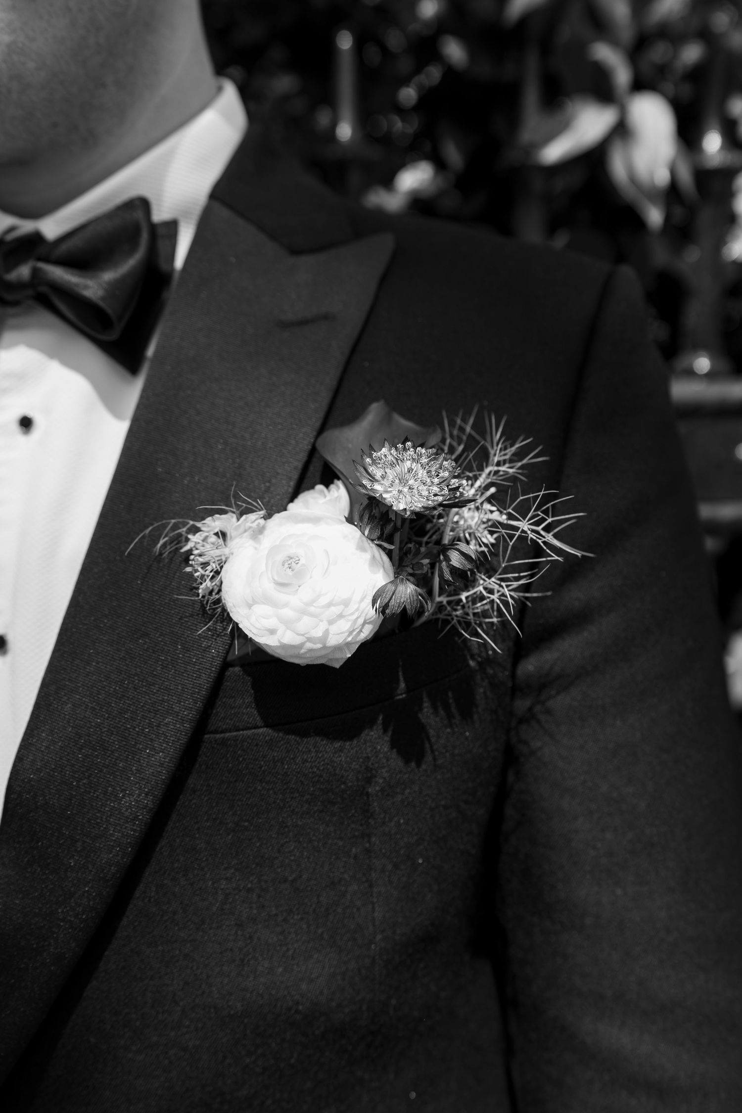 Black and white close-up of groom's boutonniere with white rose on black tuxedo lapel.