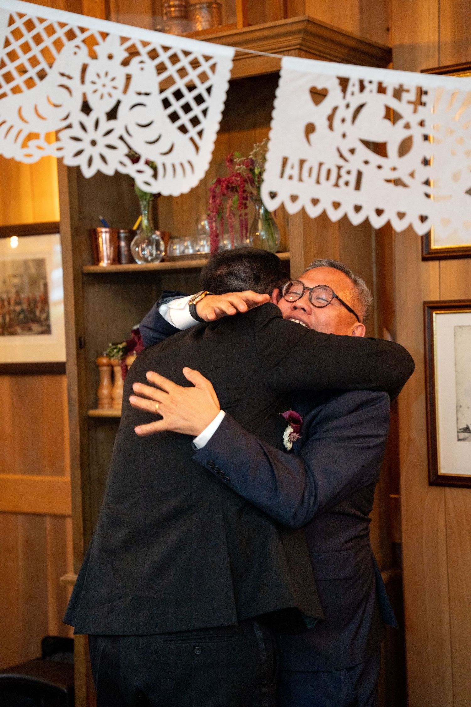 Two men share an emotional hug at a wedding reception under decorative papel picado.
