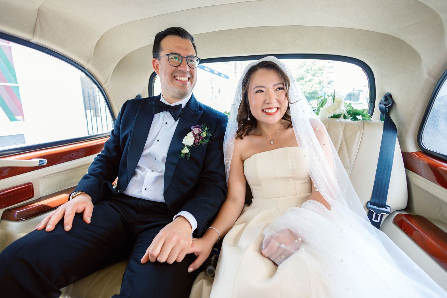 Happy bride in strapless gown and groom in navy tuxedo sitting together in a vintage wedding car.