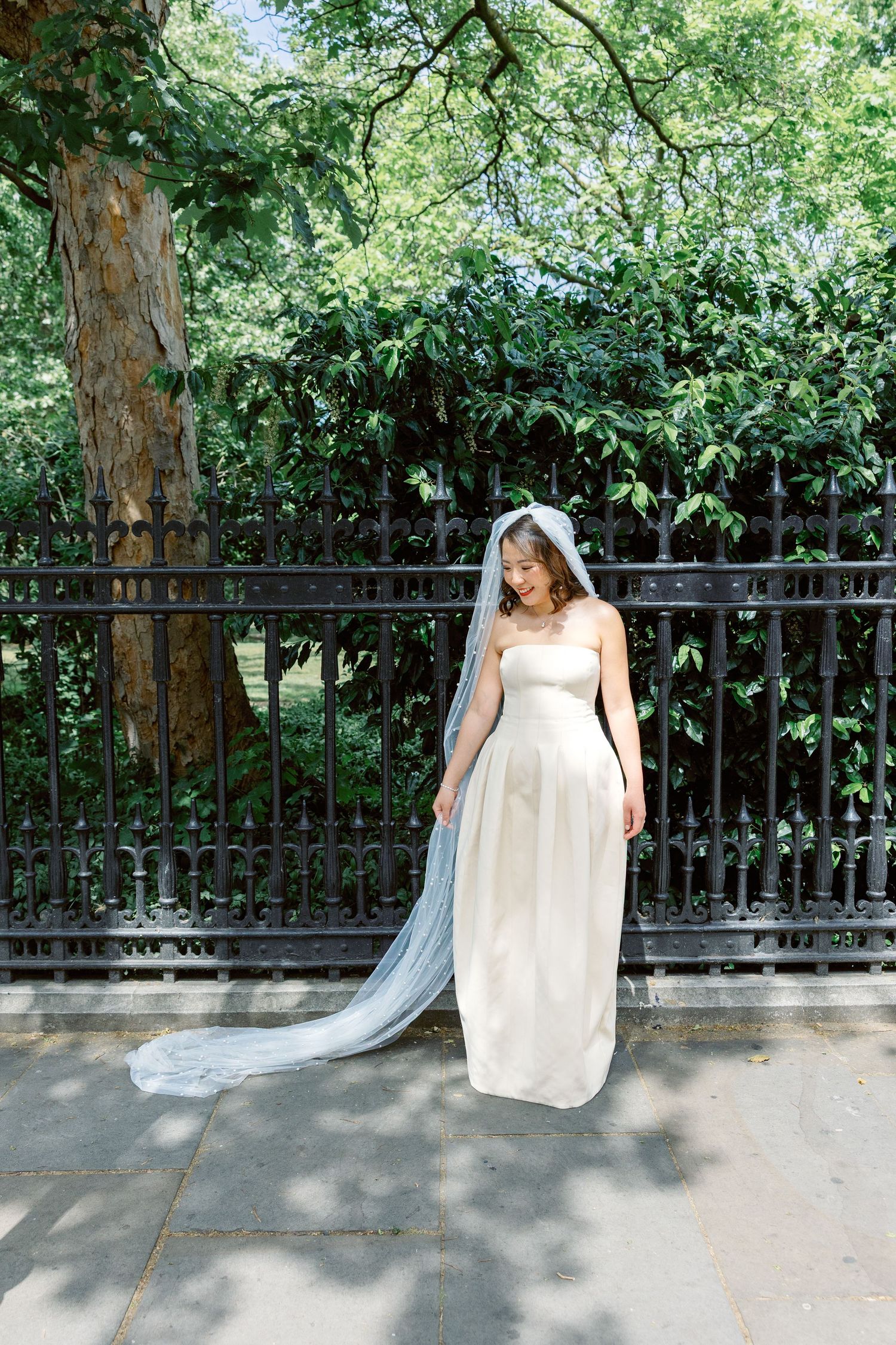 Bride in strapless white gown with long flowing veil standing by ornate iron fence in lush garden.
