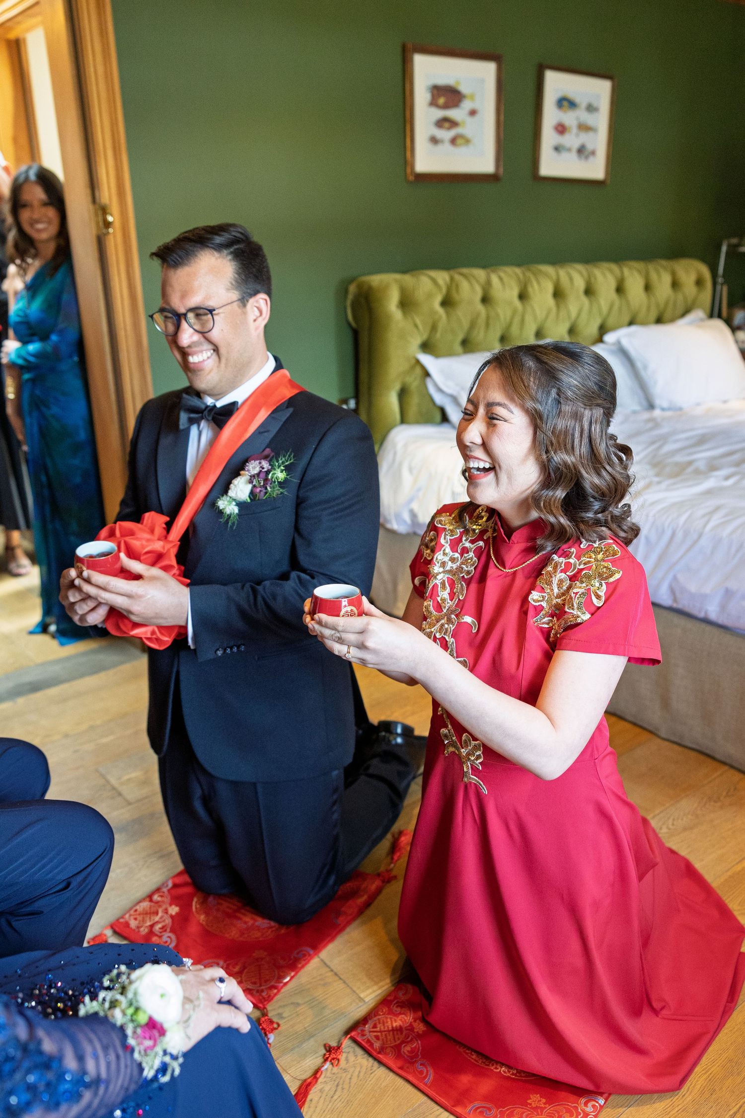 Couple in traditional Chinese tea ceremony, bride in red qipao and groom in tuxedo kneeling together.